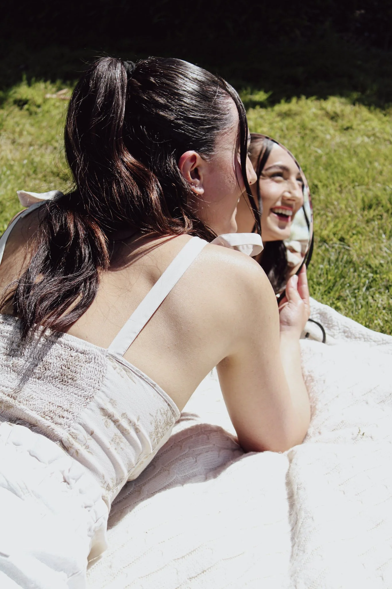 Woman with dark hair in a ponytail, smiling and holding a mirror, sitting outdoors on grass, wearing a white sleeveless top.