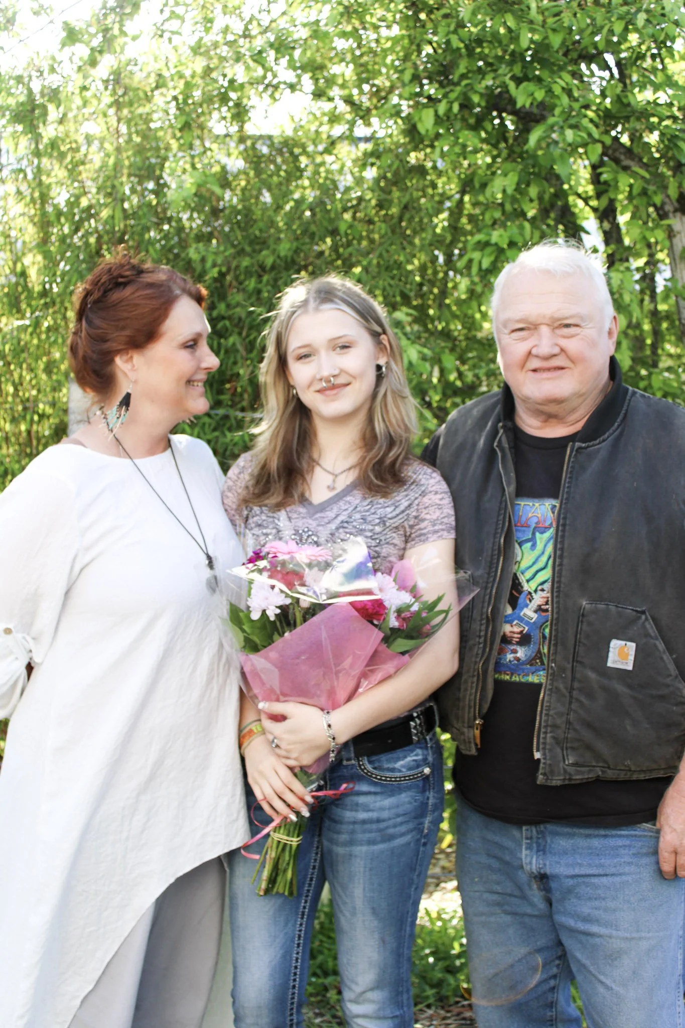 Three people standing outside in front of green trees, with the woman in the middle holding a bouquet of pink flowers. The woman on the left has short red hair, the woman in the middle has long blonde hair, and the man on the right has short white ha