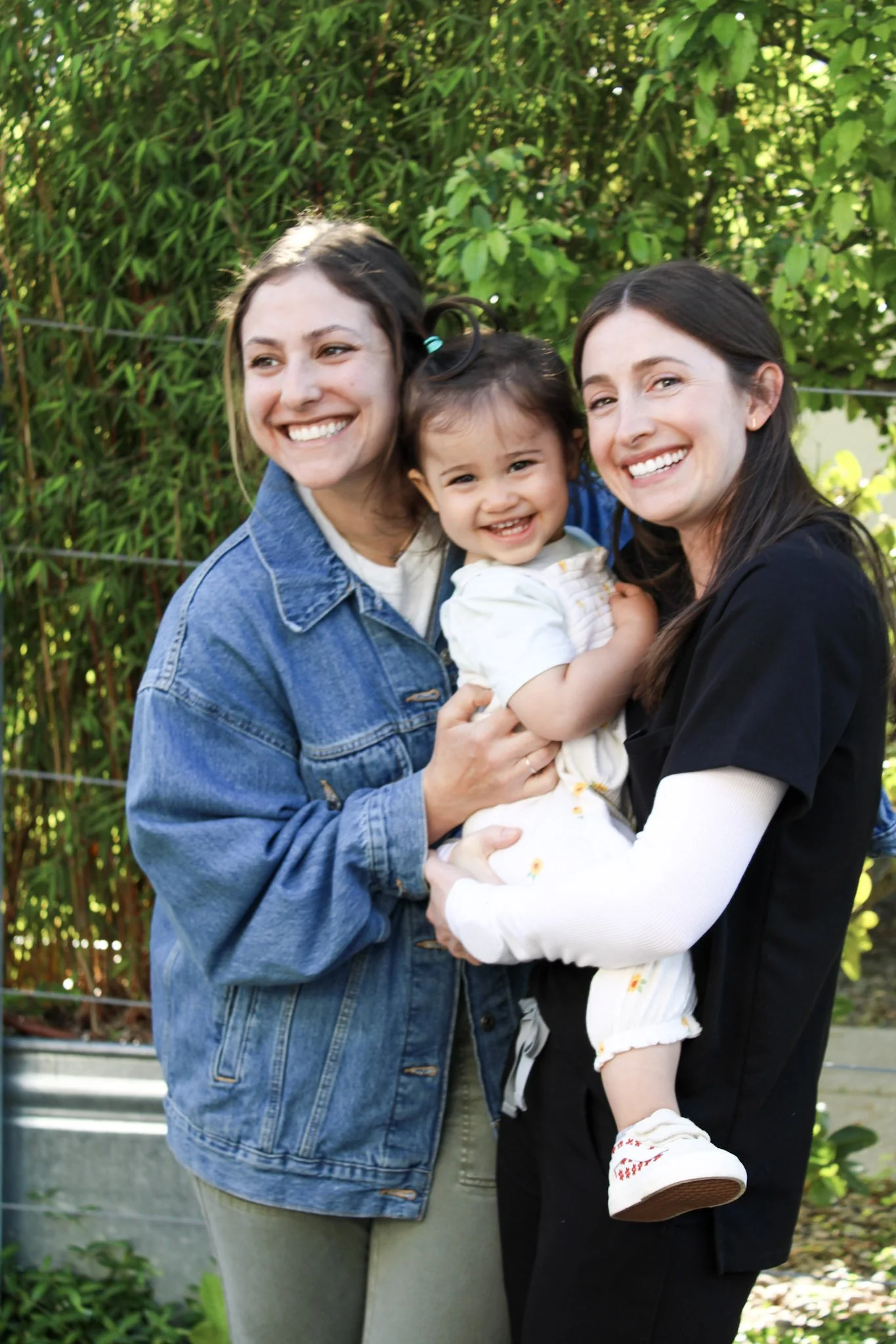 Two women and a young girl smiling outdoors with green foliage in the background, the women are holding the girl.