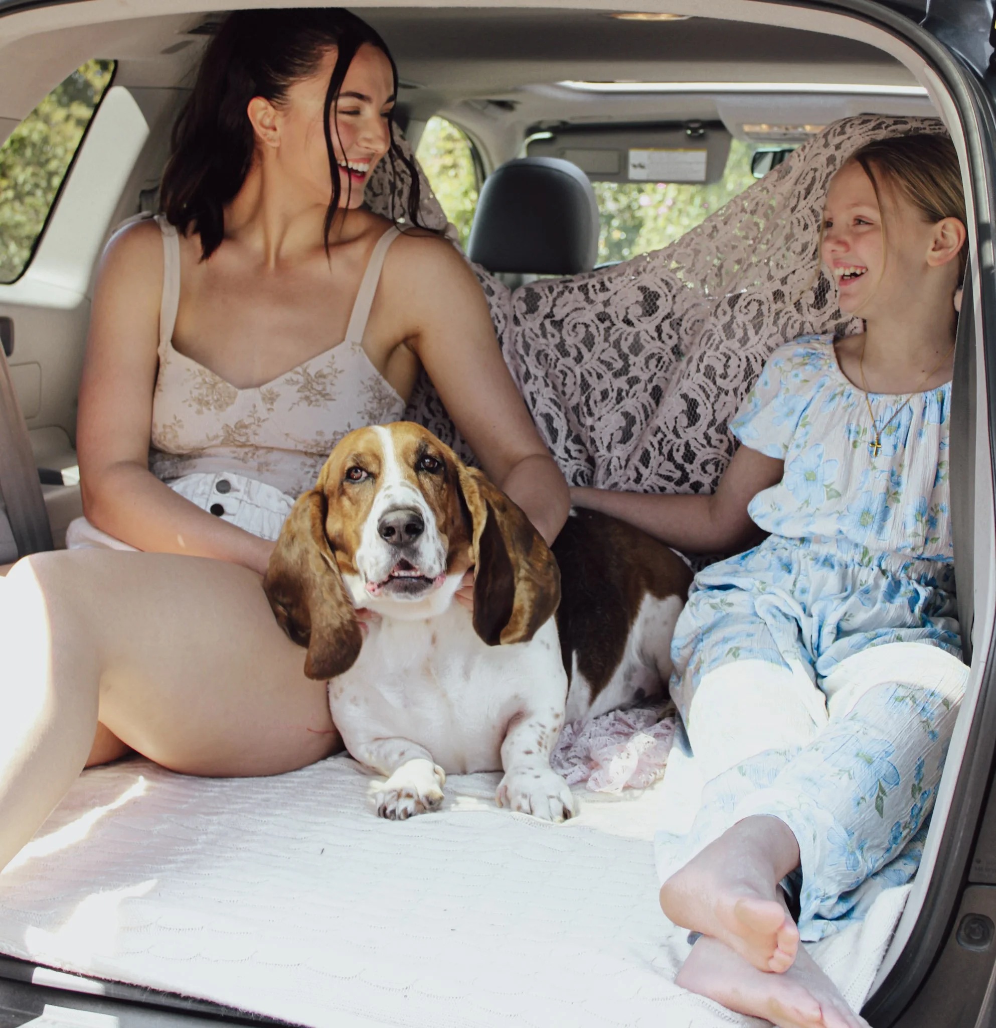 A woman and a young girl smiling and sitting in the back of a vehicle, with a dog lying on the floor in front of them. An adult's leg is visible in the foreground.