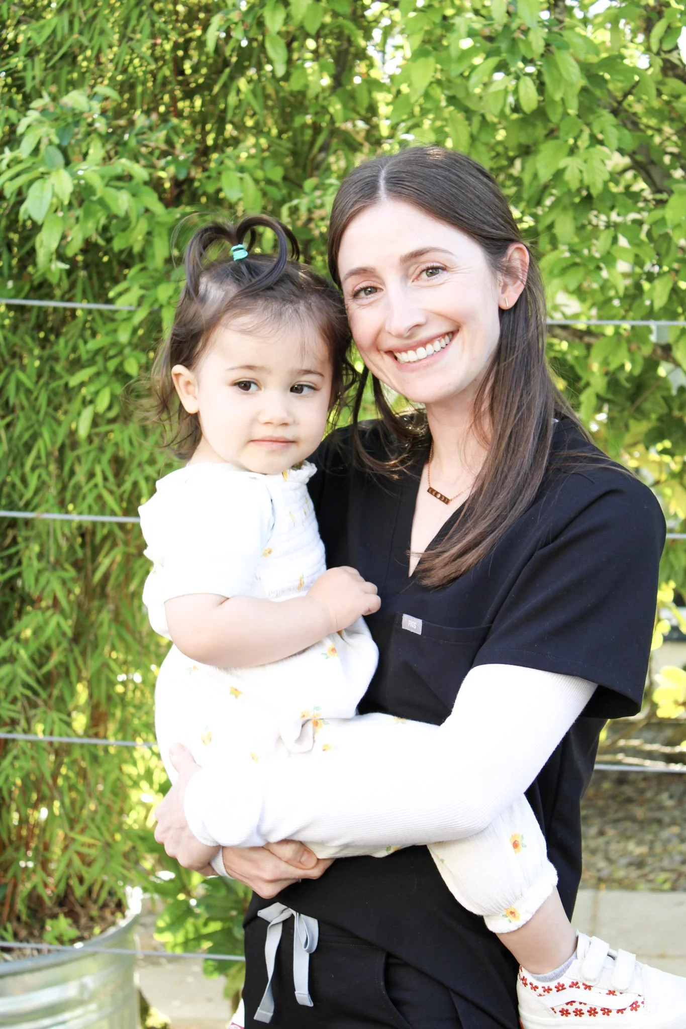 A woman with long brown hair smiling, holding a young girl with dark hair styled in pigtails, standing outdoors in front of green bushes.