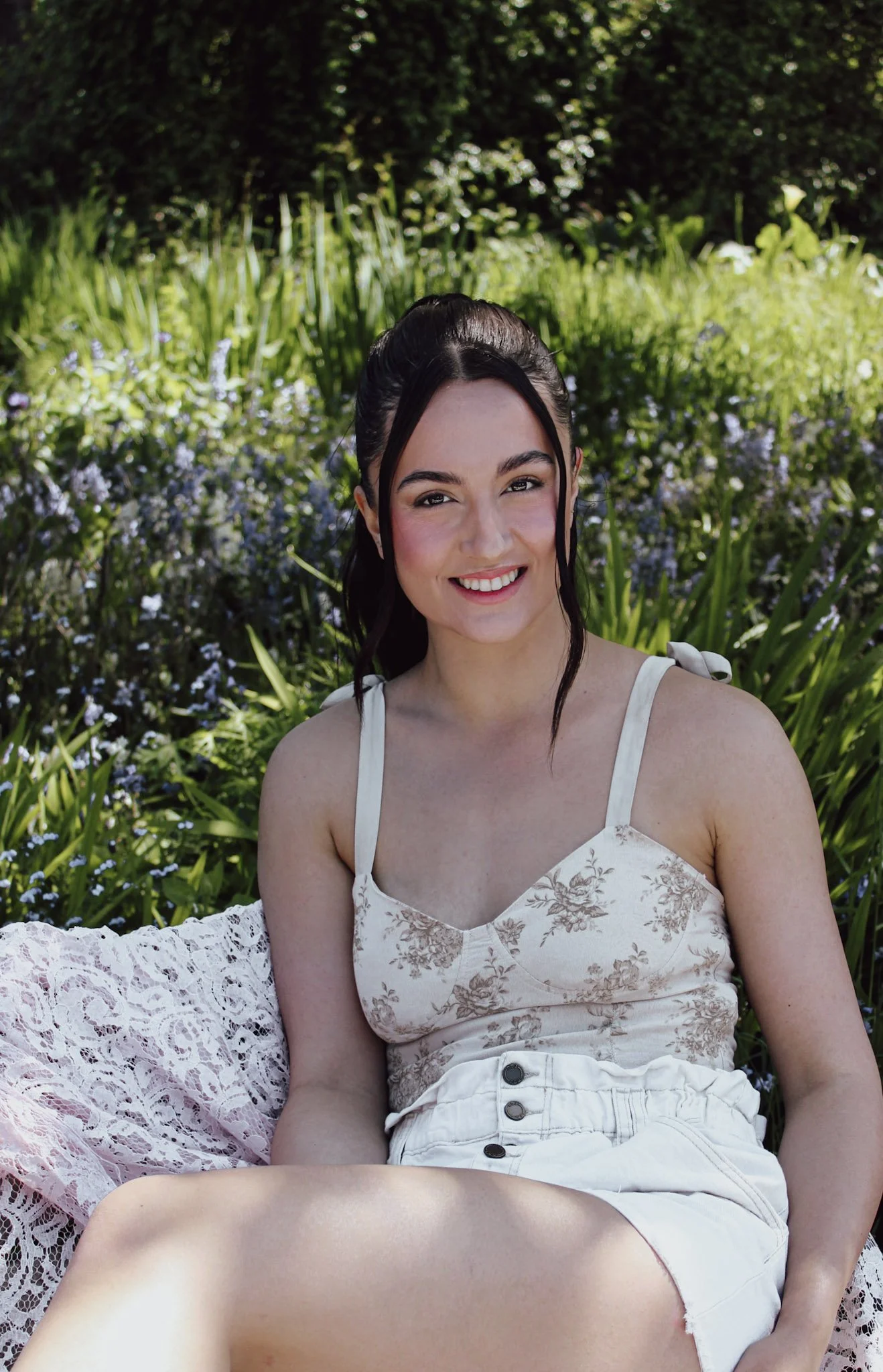 A young woman with dark hair styled in a high ponytail, sitting outdoors in front of green plants and flowers, smiling at the camera, wearing a white floral tank top and white shorts.