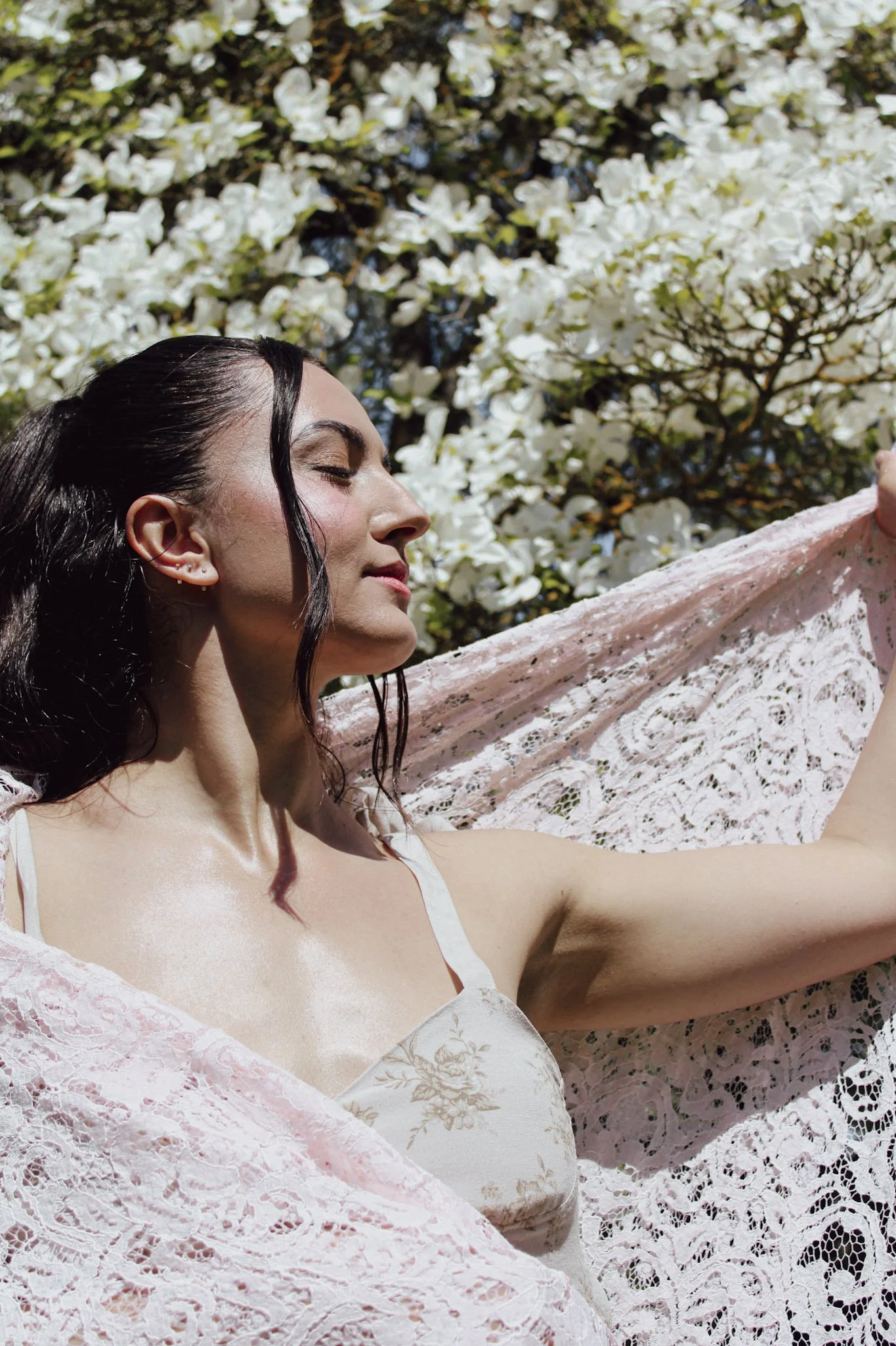 A woman with dark, wavy hair takes a selfie outdoors, wearing a white lace dress with floral details. She is standing in front of a flowering tree with white blossoms, and the sunlight highlights her face and hair.