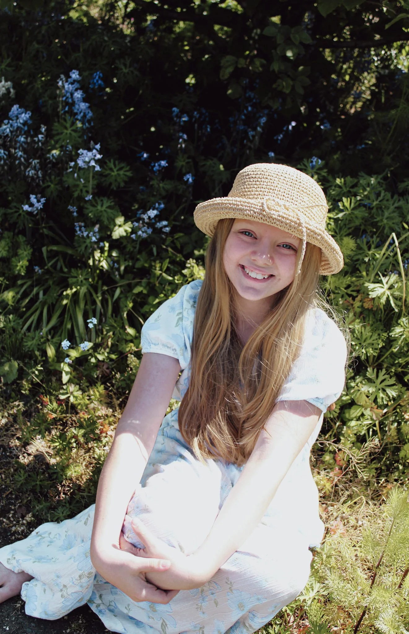 A young girl with long red hair smiling, wearing a beige wide-brimmed hat and a light-colored dress, sitting outdoors among green plants and blue flowers.