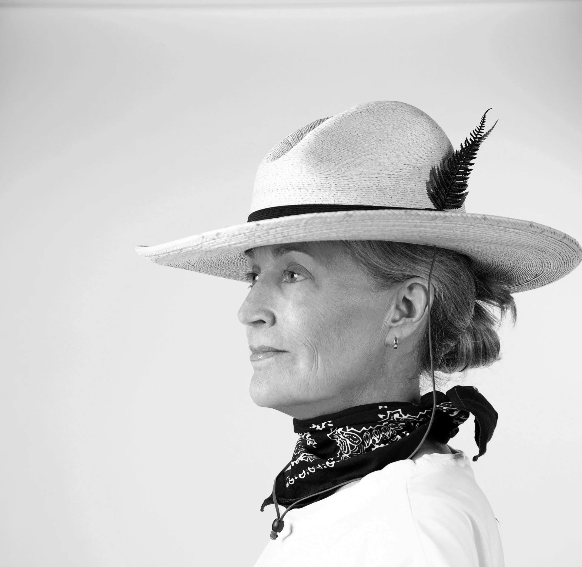 Black and white portrait of a woman wearing a wide-brimmed straw hat with black ribbon and decorative fern  on it and bandana around her neck, and a white shirt.