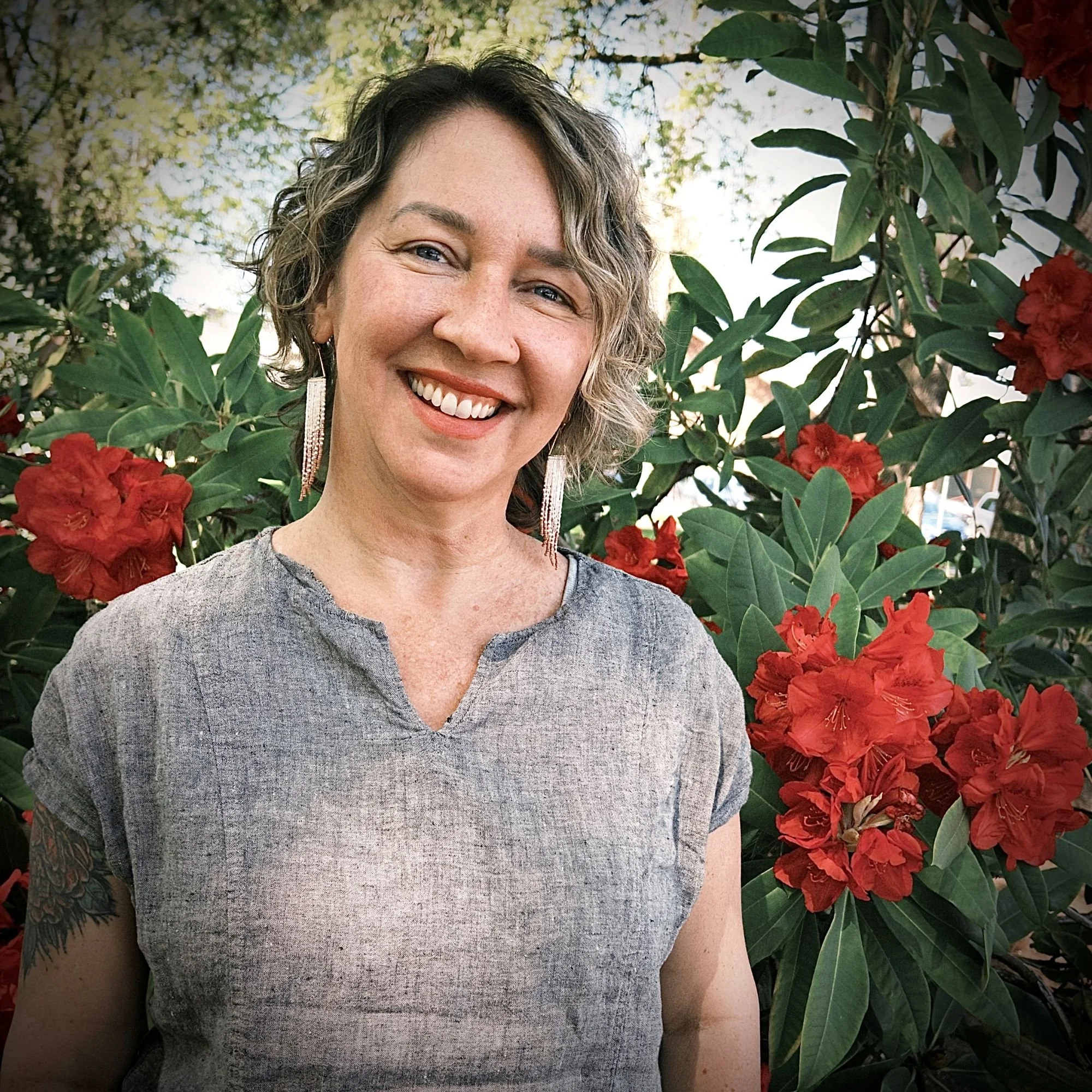 A woman with curly hair and earrings smiling in front of lush green leaves and red flowers, outdoors during the daytime.