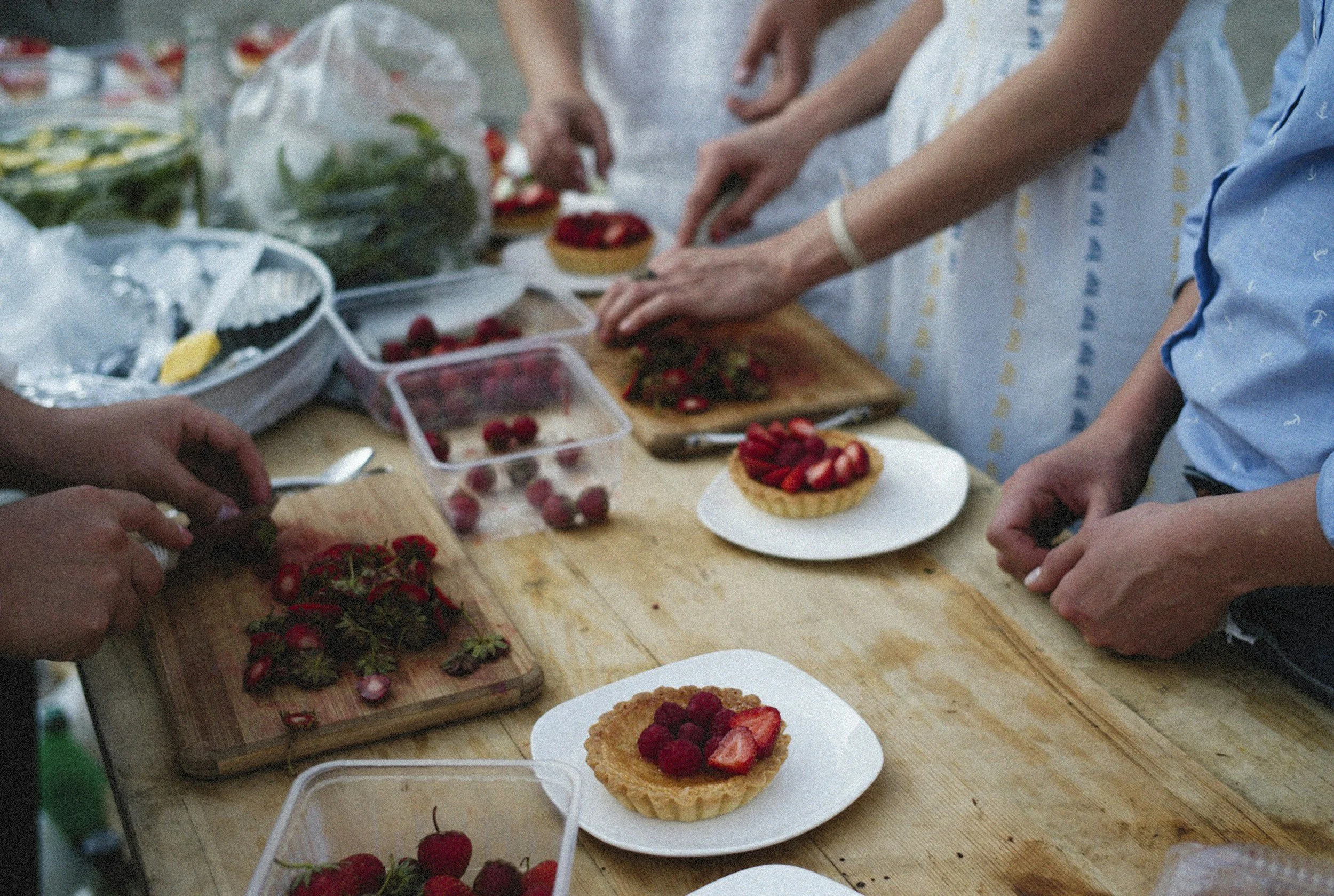 People preparing strawberry desserts with fresh strawberries, toppings, and pastry shells on a wooden table.