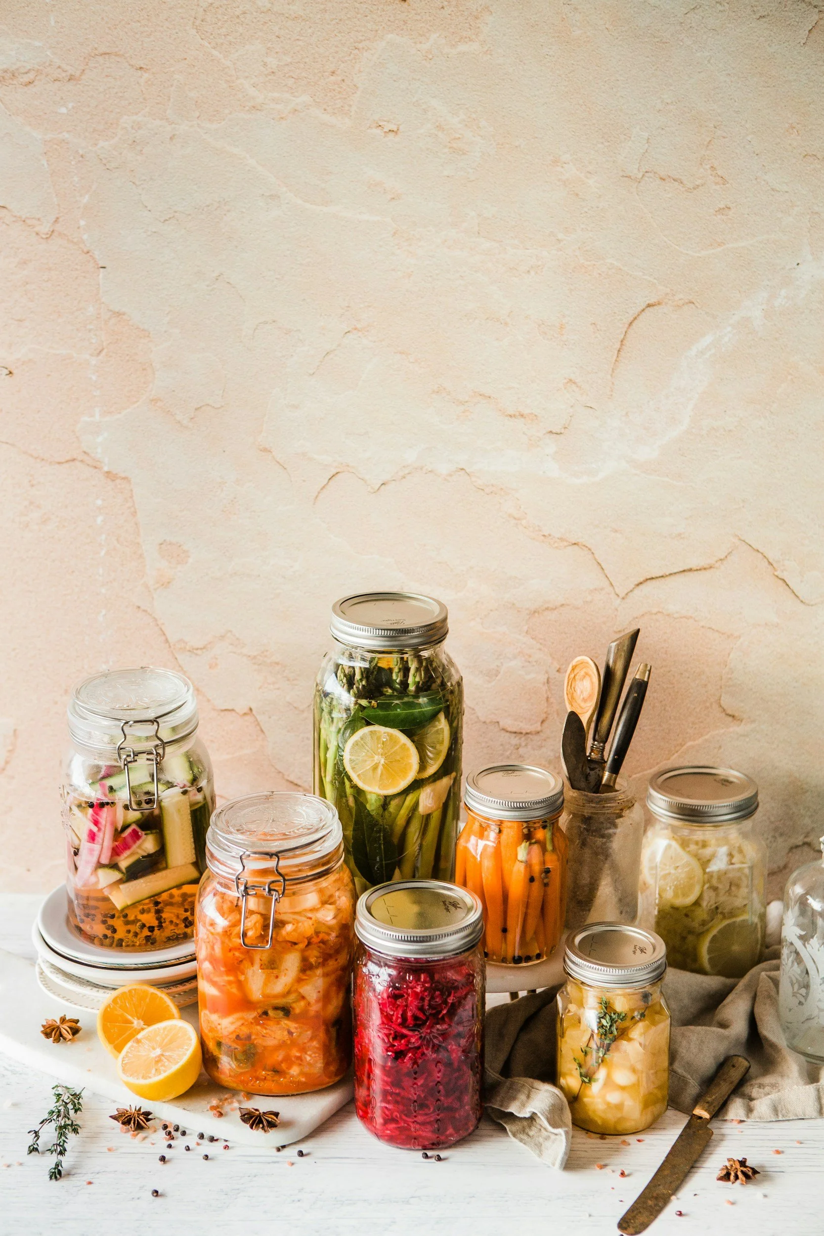 Various mason jars filled with preserved fruits and vegetables are displayed on a white table. Some jars contain lemon slices, carrots, cranberries, and herbs. There are also sliced lemons and star anise on the table. The background features a textured, light-colored wall.