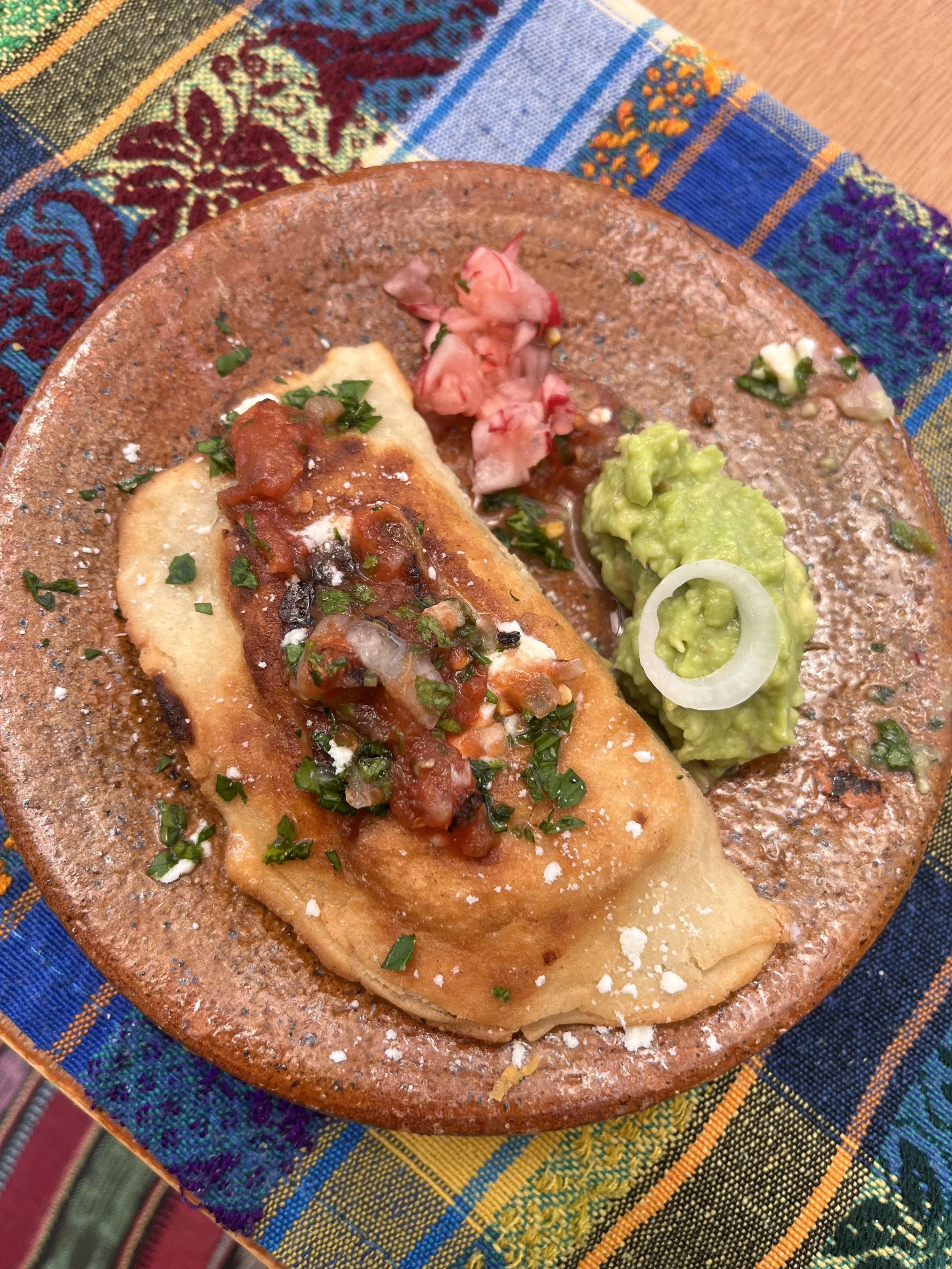 Close-up of a tostada with beans, salsa, fresh cheese, and chopped herbs, served on a rustic plate with guacamole, onions, and pickled vegetables on the side, placed on colorful, woven Guatemala fabric.