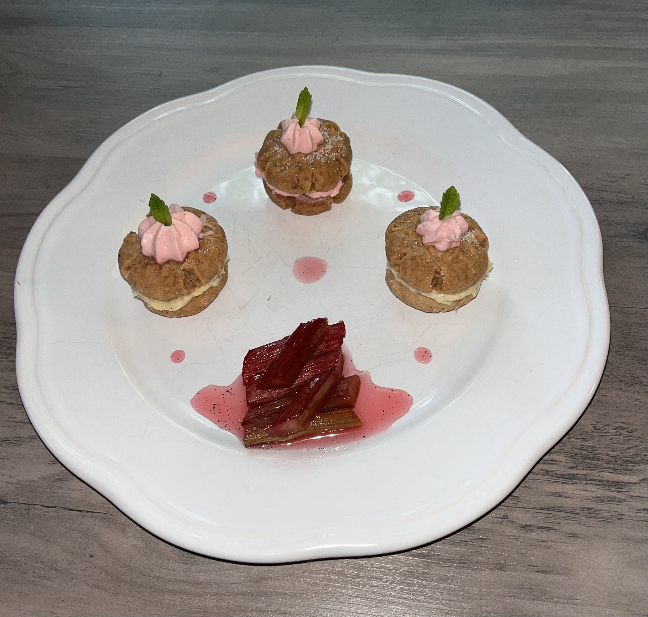 A white ceramic plate with three small dessert bites topped with pink cream and green mint leaves, garnished with red sauce drizzle on a wooden surface.