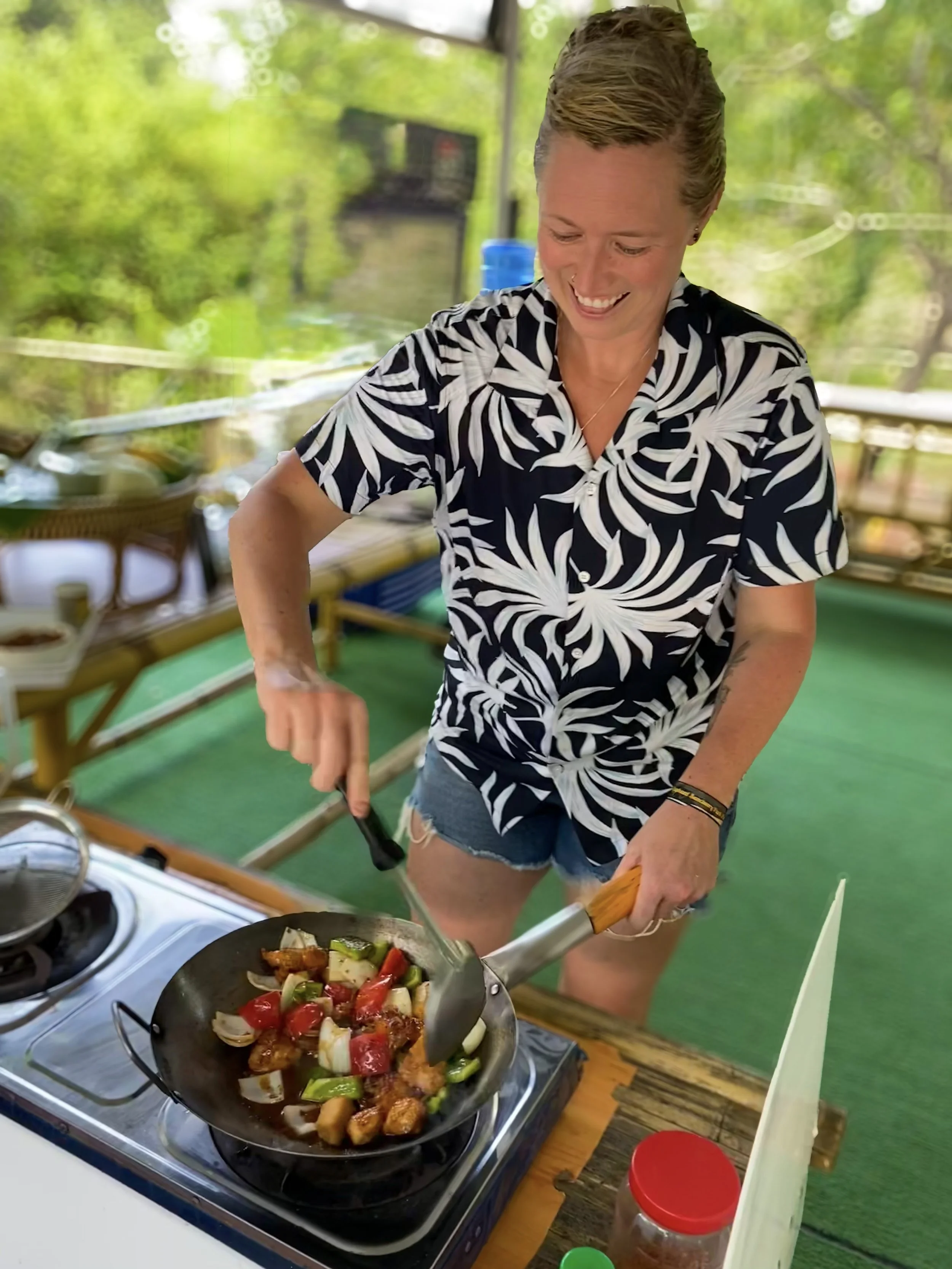 A woman wearing a black and white patterned shirt and denim shorts cooking vegetables in a frying pan on an outdoor stove, smiling and looking down, with a blurred green trees background.