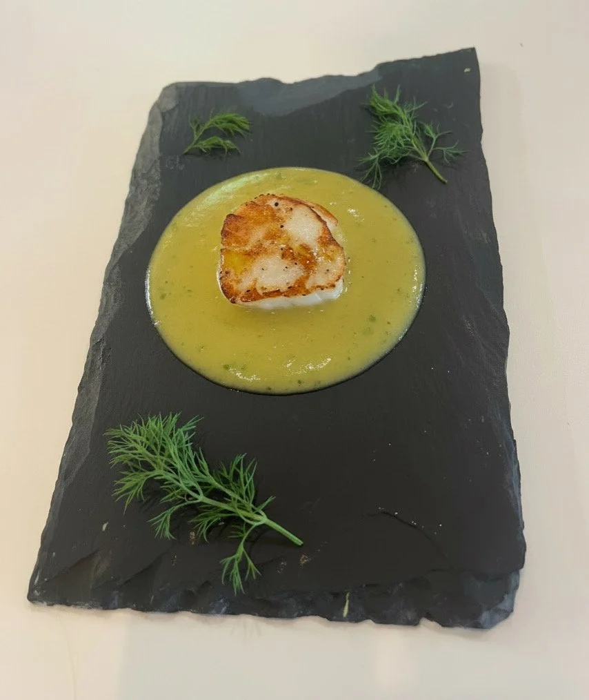 Seared scallop on a bed of green sauce, garnished with fresh herbs on a black slate serving platter.