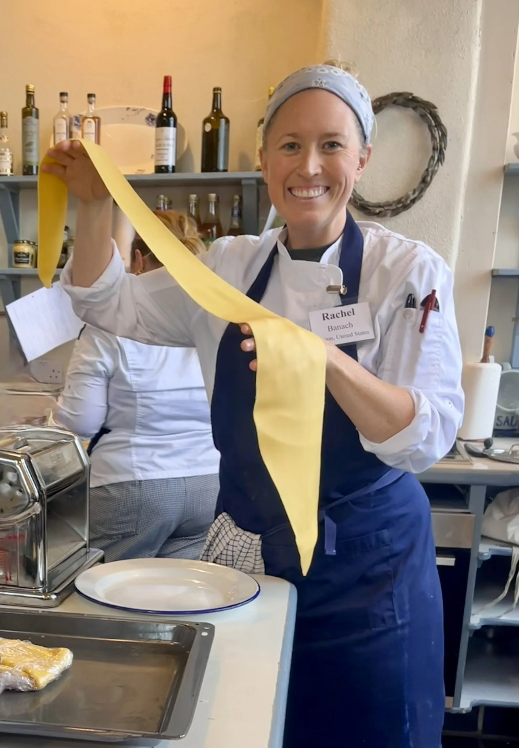 Dr. Rachel Banach is smiling and holding a length of fresh made rolled yellow pasta in a kitchen. She is wearing a chef's coat, apron, and headscarf.