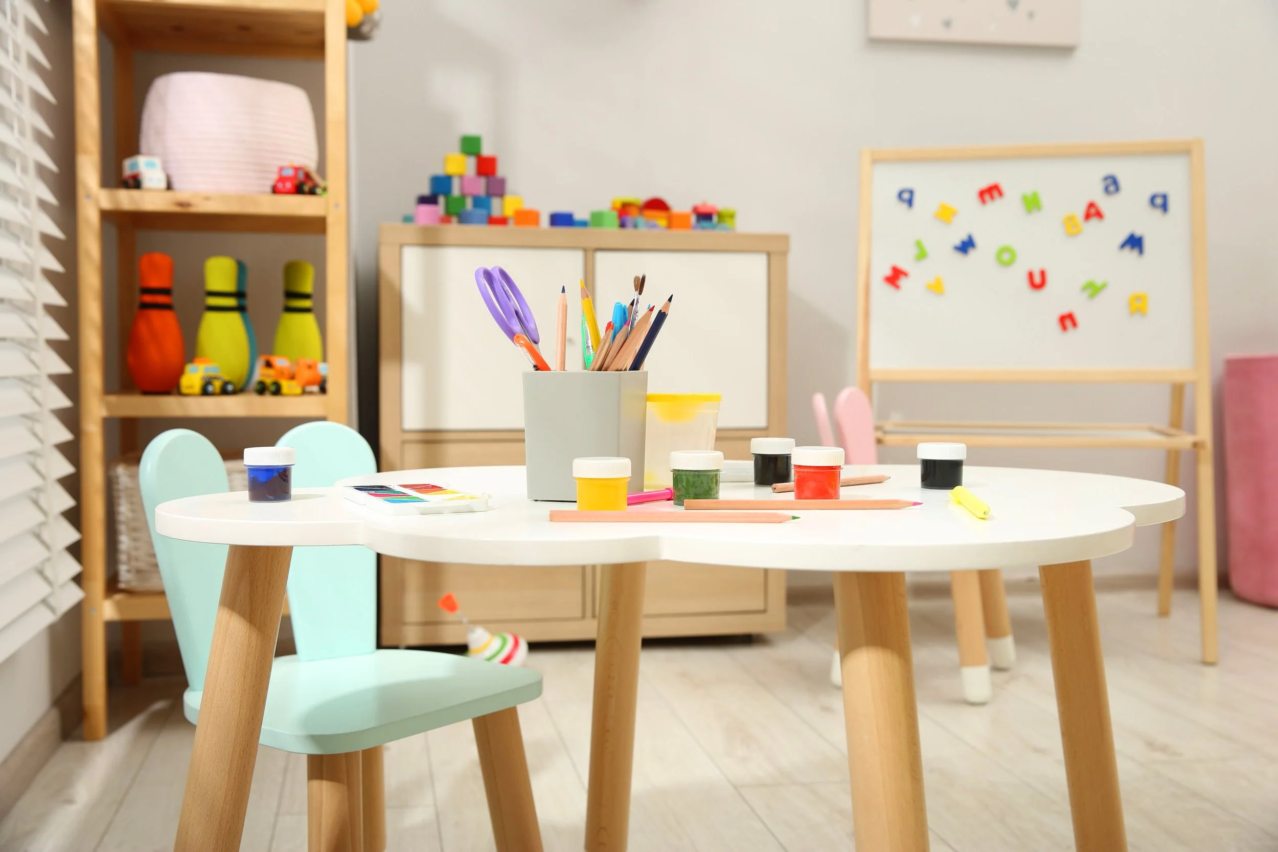 A children's craft room with a white circular table holding art supplies like colored paints, markers, brushes, and pencils. Light green chairs are around the table. In the background, shelves hold toy bowling pins, toy cars, and colorful building blocks. A whiteboard with colorful alphabet letters and shapes is on the wall.