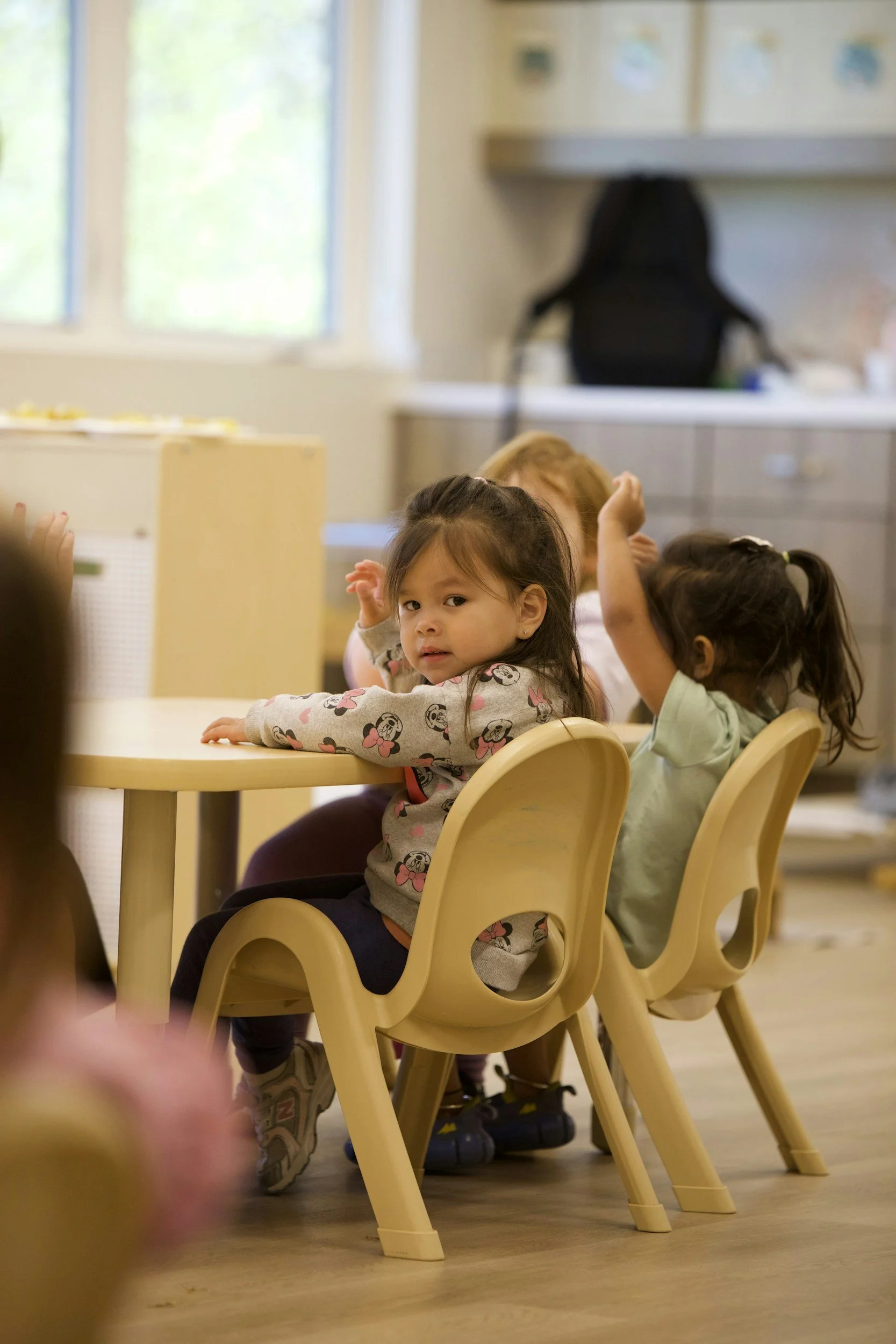 Young children sitting at a yellow table in a classroom, with some looking directly at the camera and others engaging in activities; background includes a window with natural light.