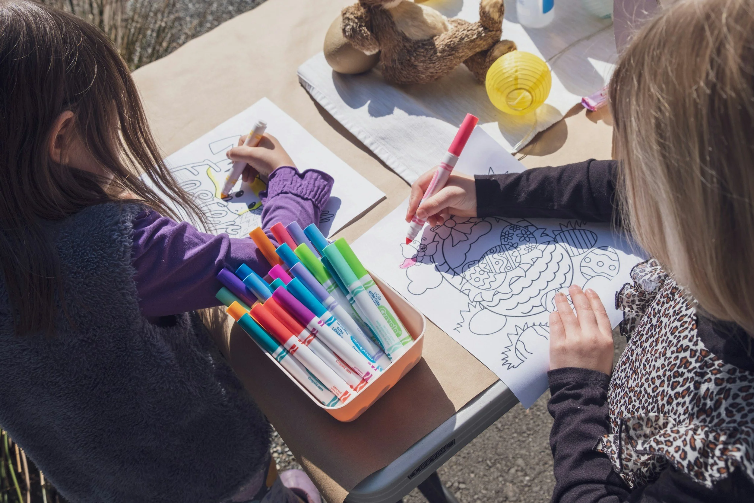 Two children sketching and coloring on paper at an outdoor table with colorful markers, with decorative objects including a yellow paper lantern and a wooden sculpture nearby.