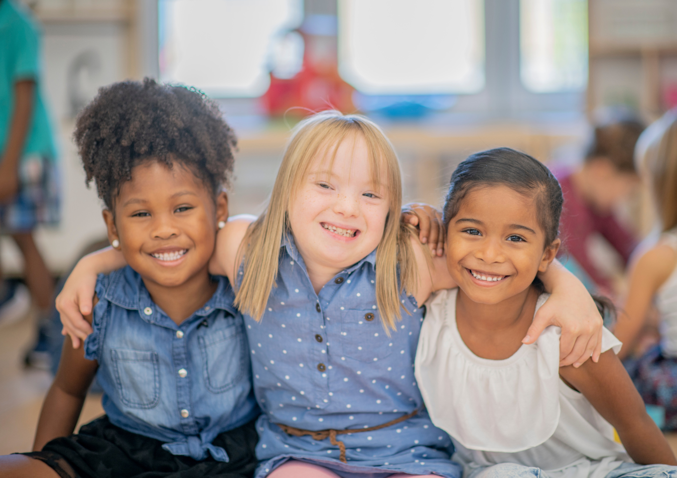 Three smiling young girls sitting close together in a classroom, with arms around each other.