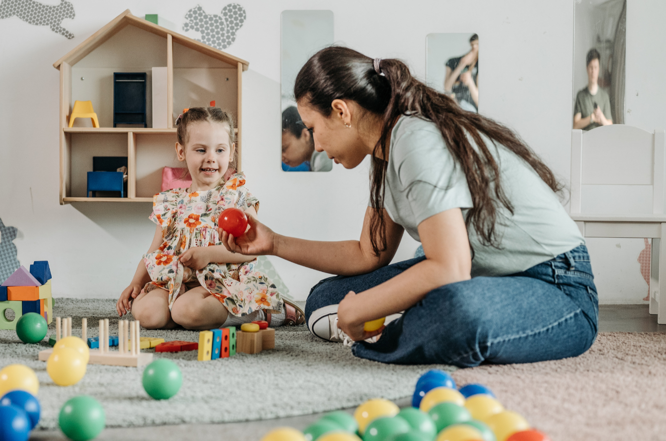 A woman and a young girl playing with colorful balls and toys on a carpeted floor in a playroom.