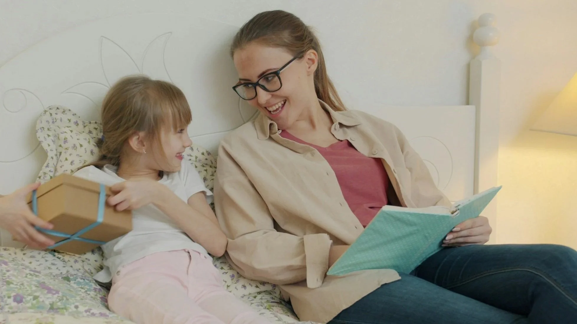 A woman and a young girl sitting on a bed. The woman is reading a book to the girl, who holds a small gift wrapped with a blue ribbon. They are smiling and looking at each other, sharing a joyful moment.