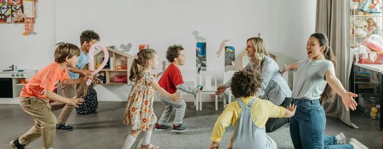 Children and two women playing and dancing in a playroom with toys and toys on shelves.