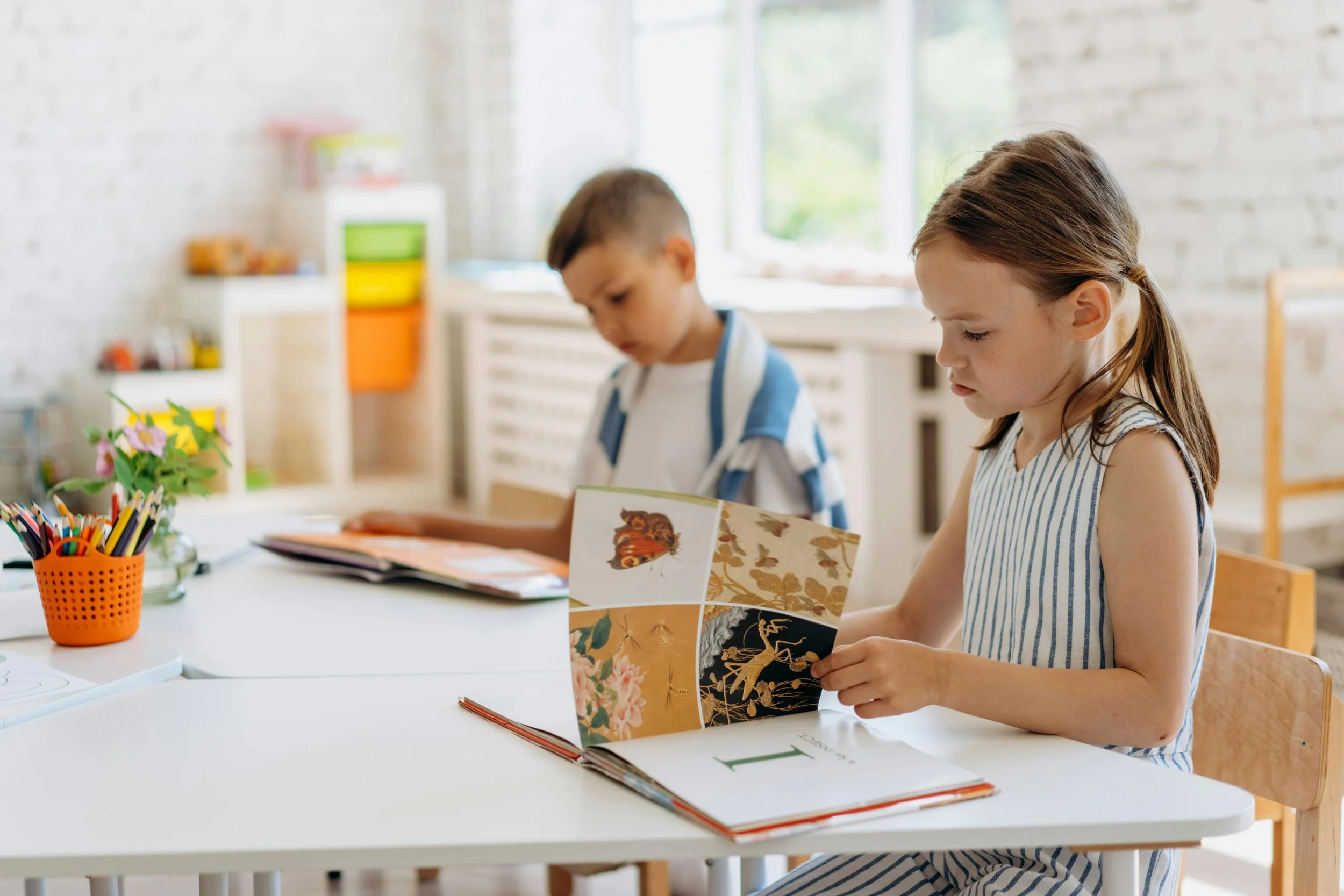 Two children, a boy and a girl, sitting at a white table in a bright room. The girl is reading a colorful book, and the boy is looking down at something on the table. There is an orange container with colored pencils and a small potted plant on the table.