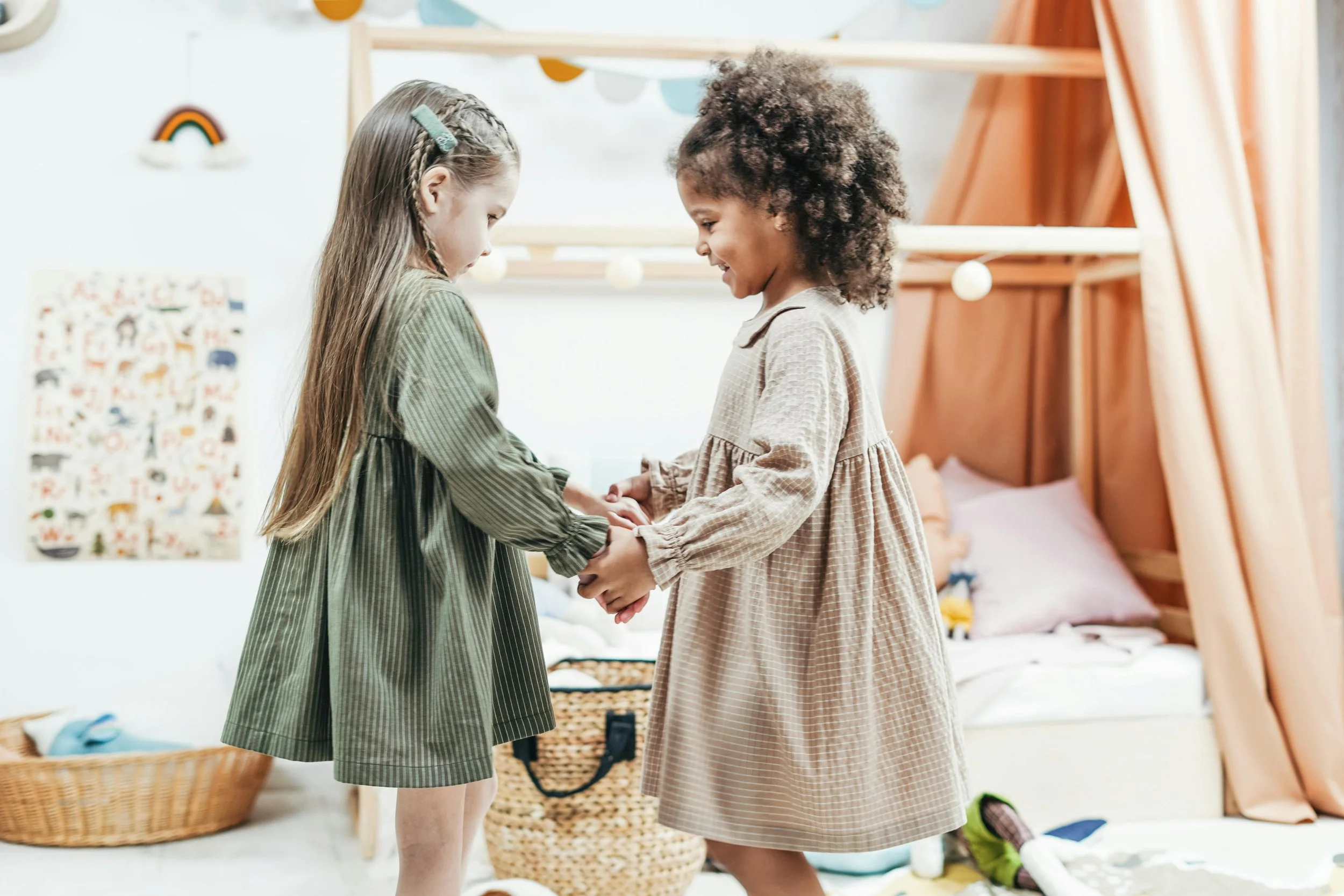 Two young girls are holding hands and smiling at each other in a playroom. One girl has long brown hair styled in braids with a clip, and the other has curly hair. The room has a bed with pink curtains, and colorful wall decorations, including a rainbow and animal pictures.