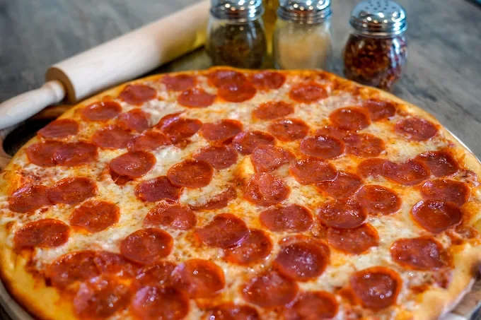 Pepperoni pizza on a wooden table with a rolling pin and spice jars in the background.