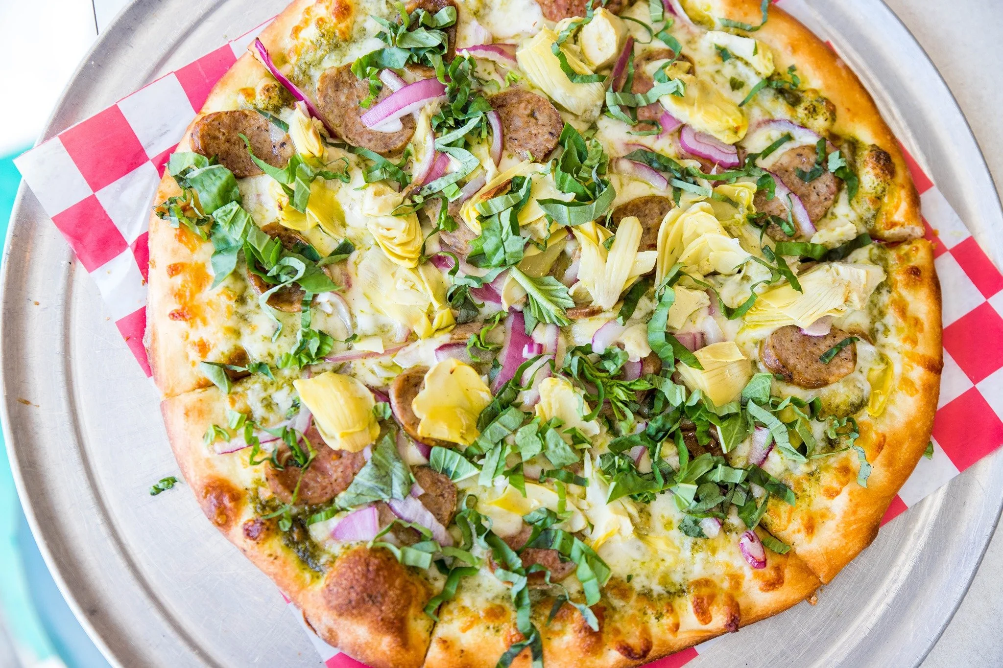 A pizza with sausage slices, artichoke hearts, red onions, melted cheese, and chopped green herbs on a metal tray lined with checkered paper.