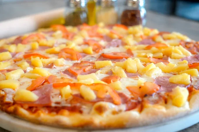 Close-up of a pineapple and ham pizza topped with sliced peppers on a pizza pan with bottles of condiments in the background.