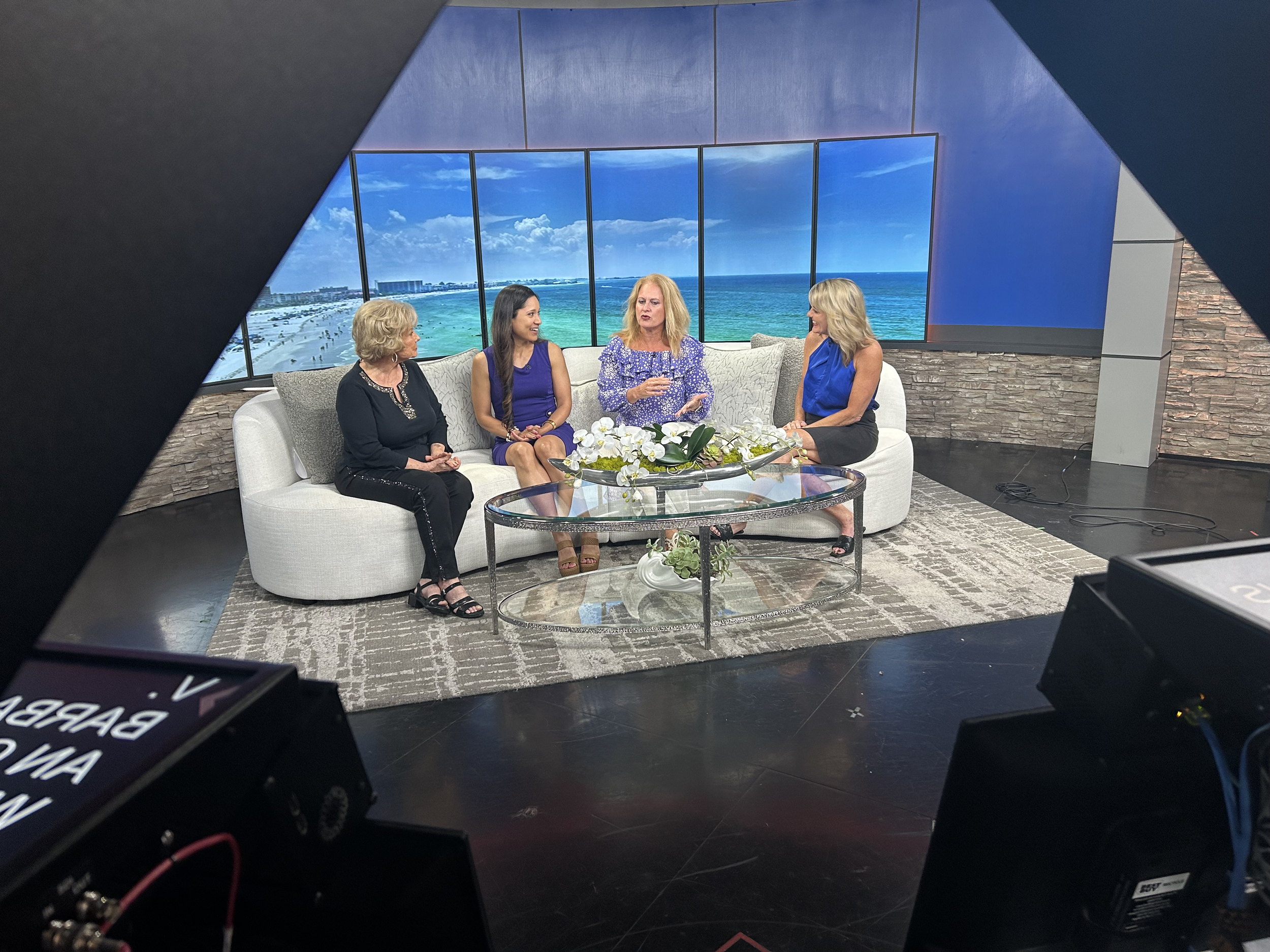 Four women sitting on a white sofa having a conversation in a TV studio with a large virtual window showing a beach scene.