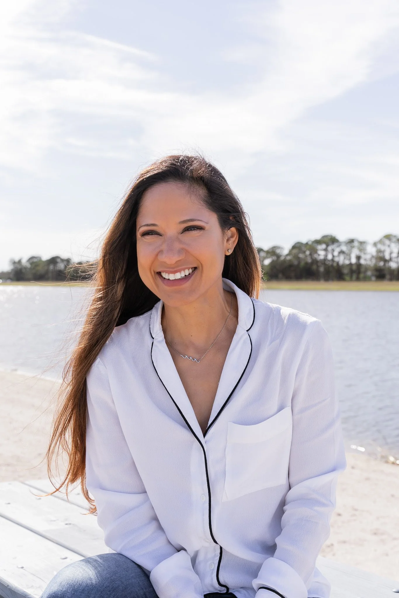 A woman sitting outdoors by a body of water, smiling, with trees in the background and a partly cloudy sky.