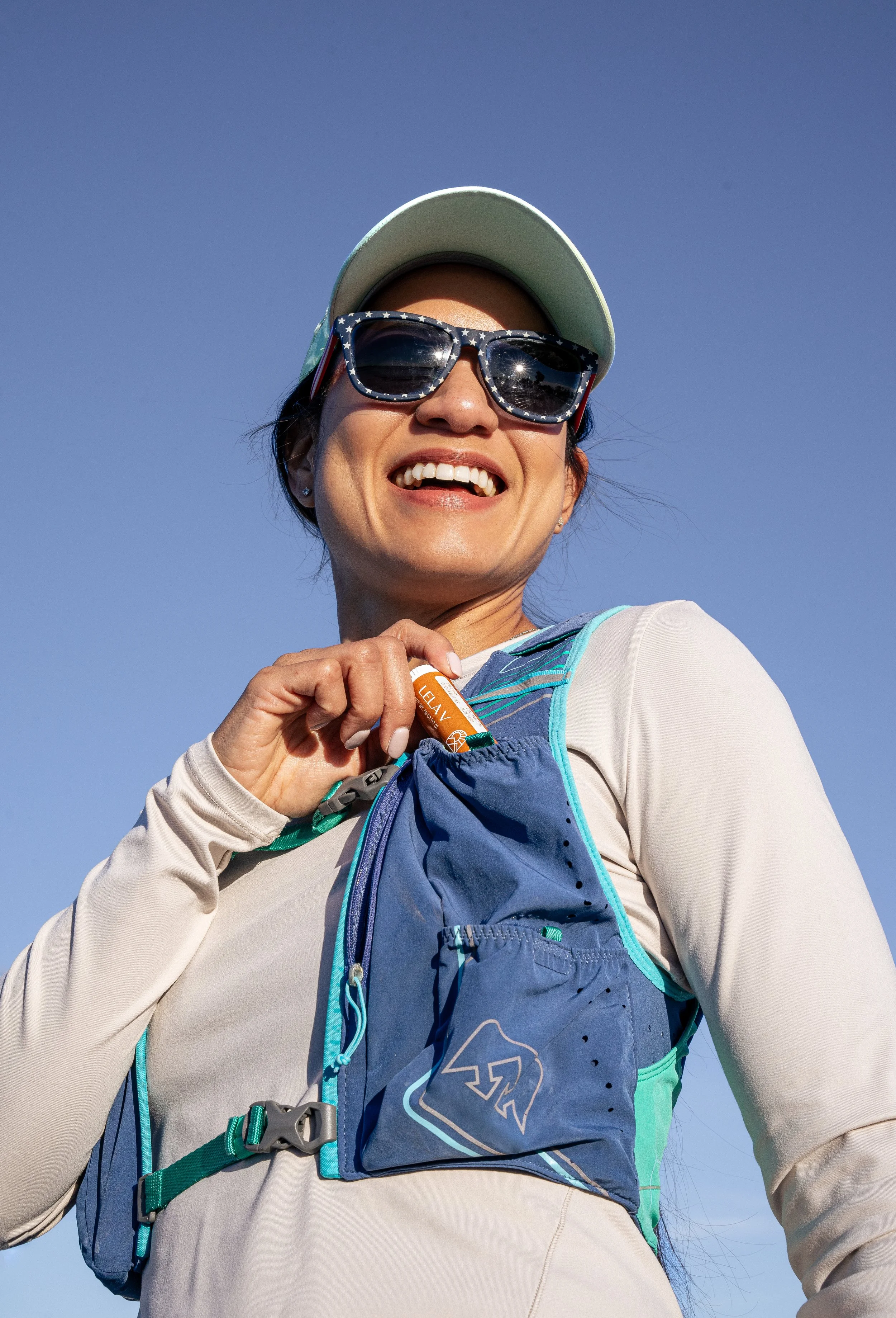 Close-up of a smiling woman wearing sunglasses, a light-colored cap, and a lightweight running shirt, outdoors against a clear blue sky, carrying a hydration pack.