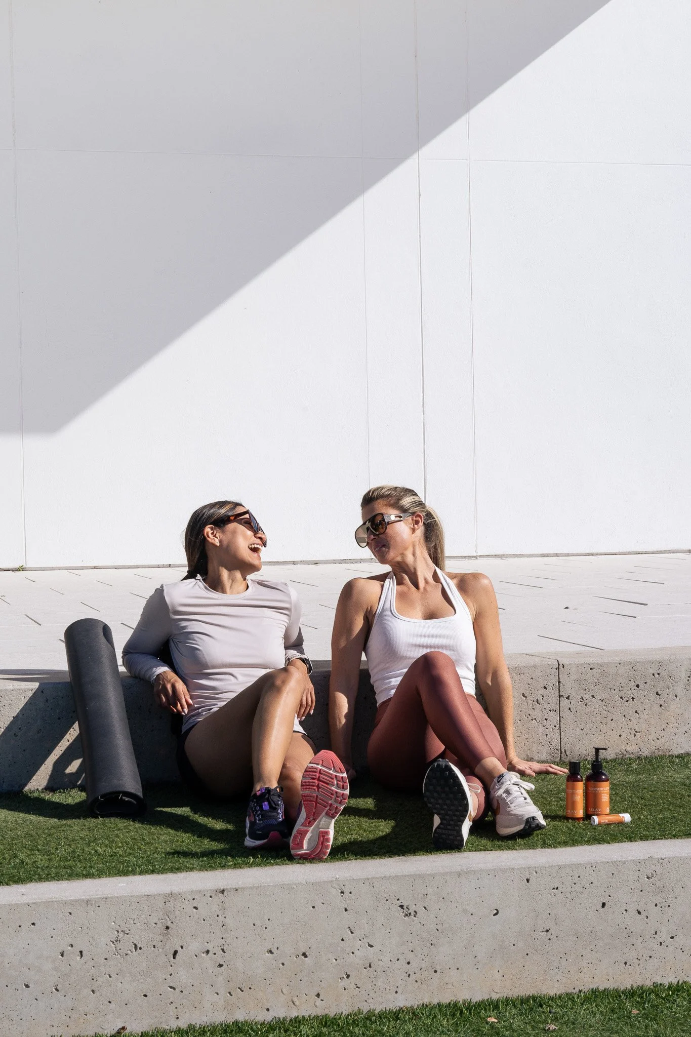 Two women sitting on a concrete ledge outdoors, smiling and talking, with workout bottles and a foam roller nearby, against a plain white wall and partly sunny sky.