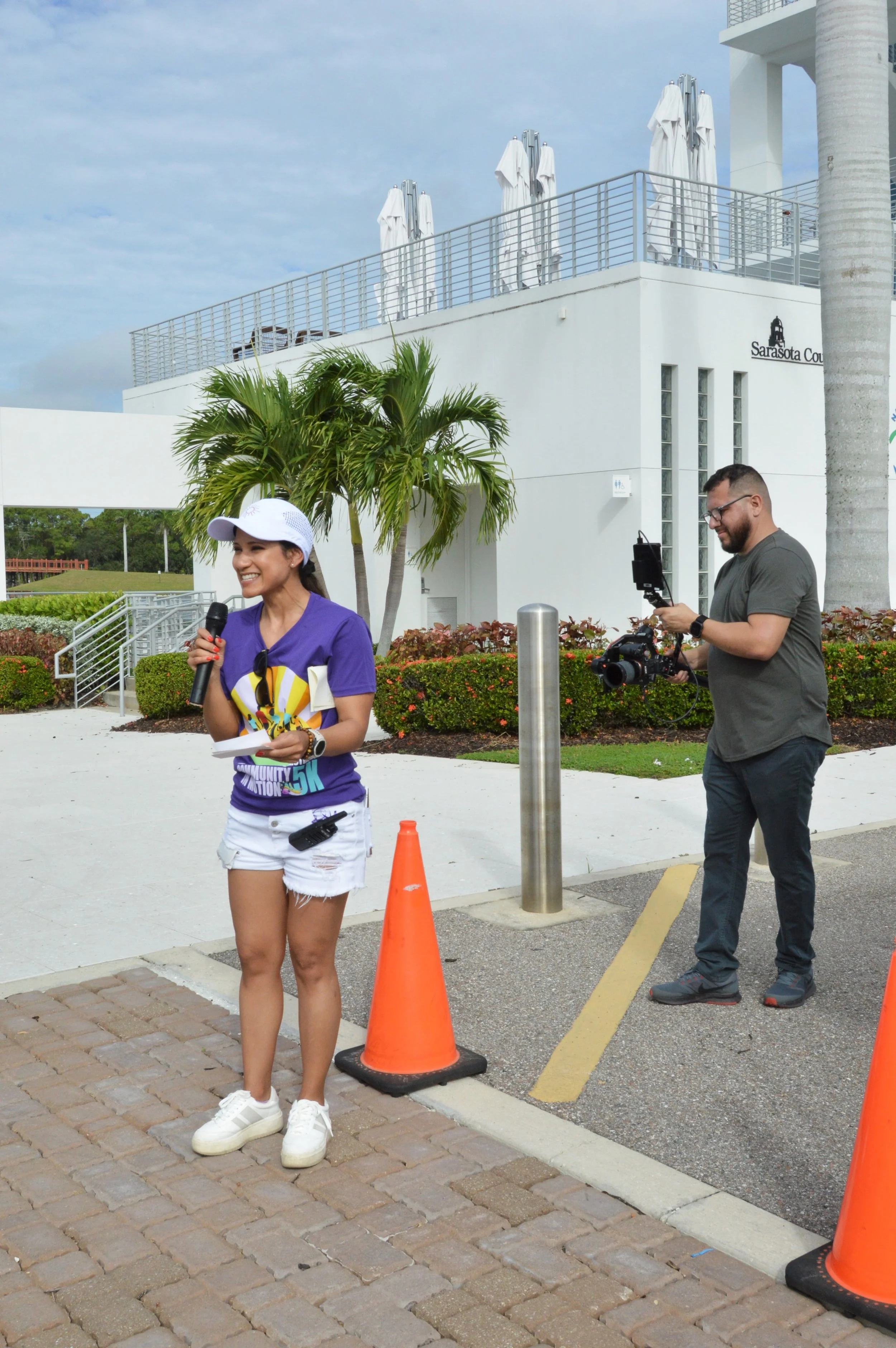 A woman wearing a purple t-shirt, white shorts, white sneakers, and a white cap holds a microphone, standing outdoors on a brick sidewalk, with a man in a gray shirt and pants operating a camera behind her. There are orange traffic cones and a palm tree nearby, and a modern white building in the background.