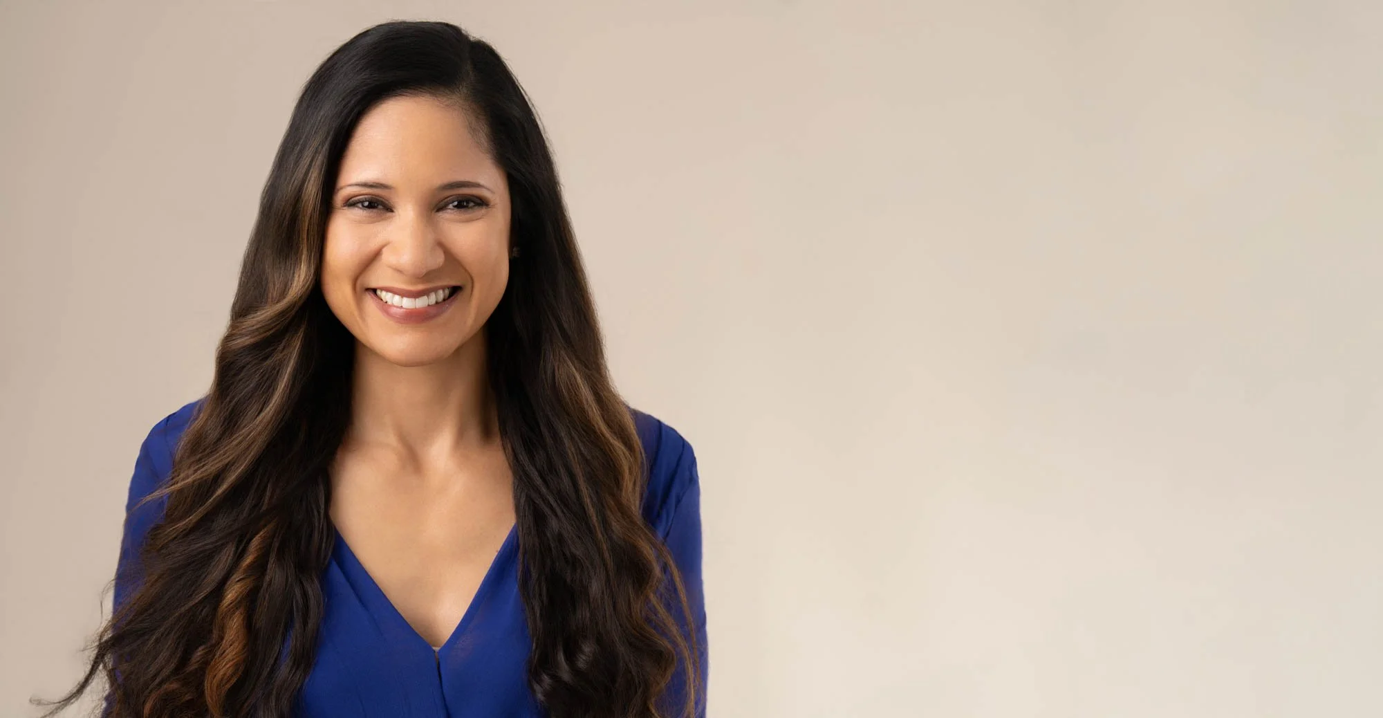 Smiling woman with long wavy dark hair wearing a blue blouse, standing against a plain beige background.