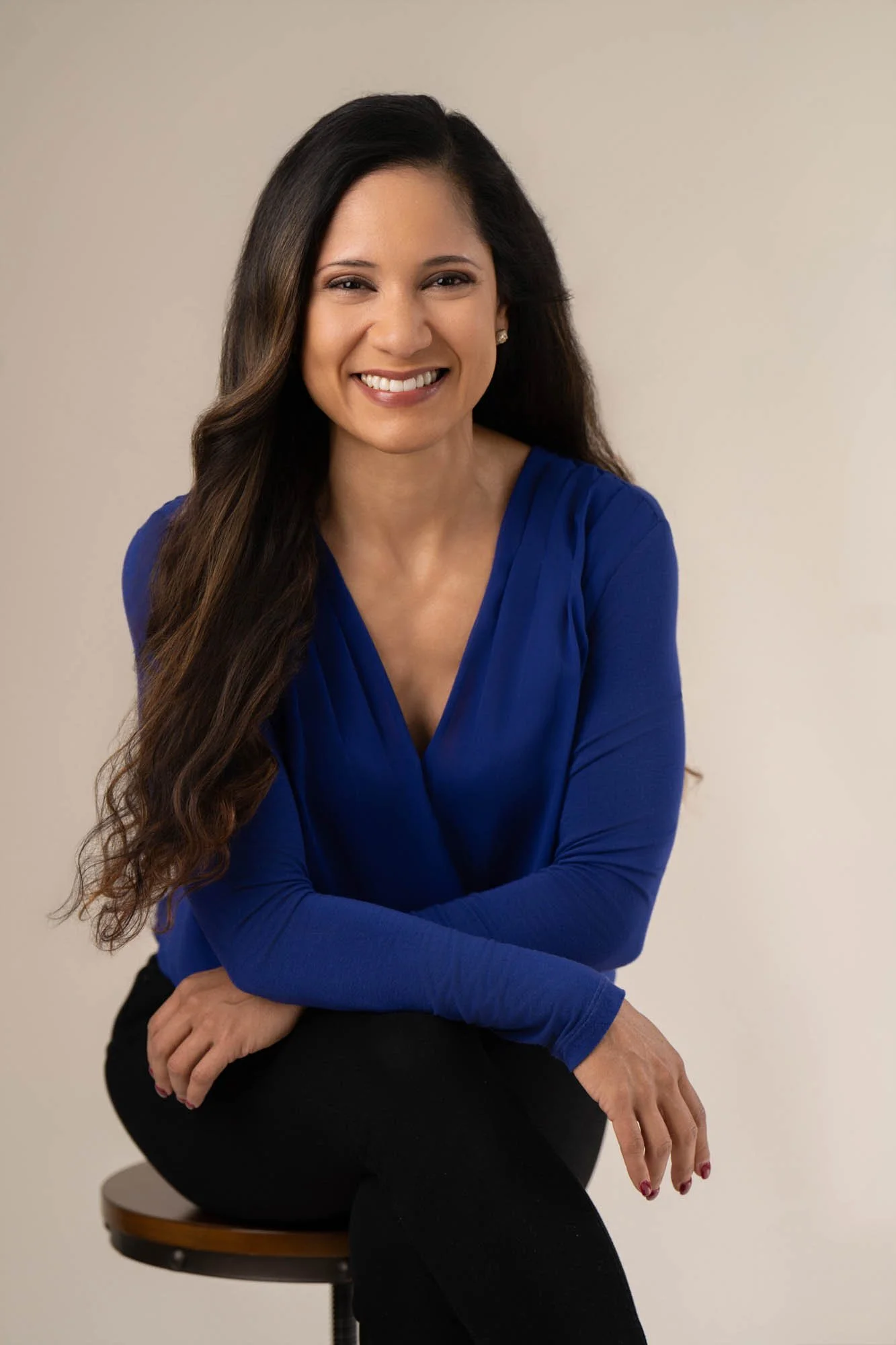 A smiling woman sitting on a stool, wearing a royal blue blouse and black pants, with long wavy hair, against a plain light-colored background.