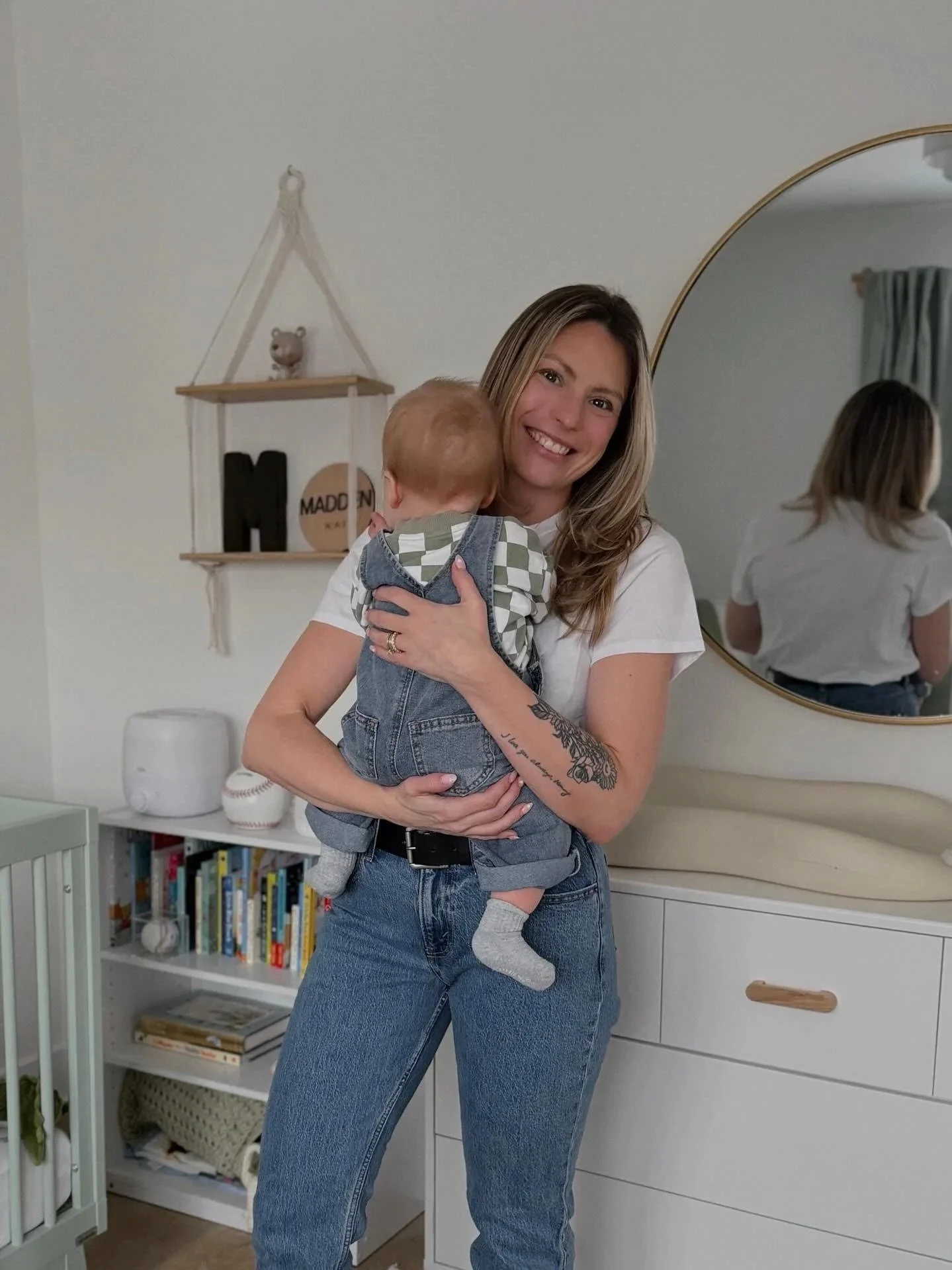A smiling woman with long, wavy hair holding a small child with red hair in her arms. They are in a cozy, well-decorated room with a mirror, bookshelf, and wall shelves.