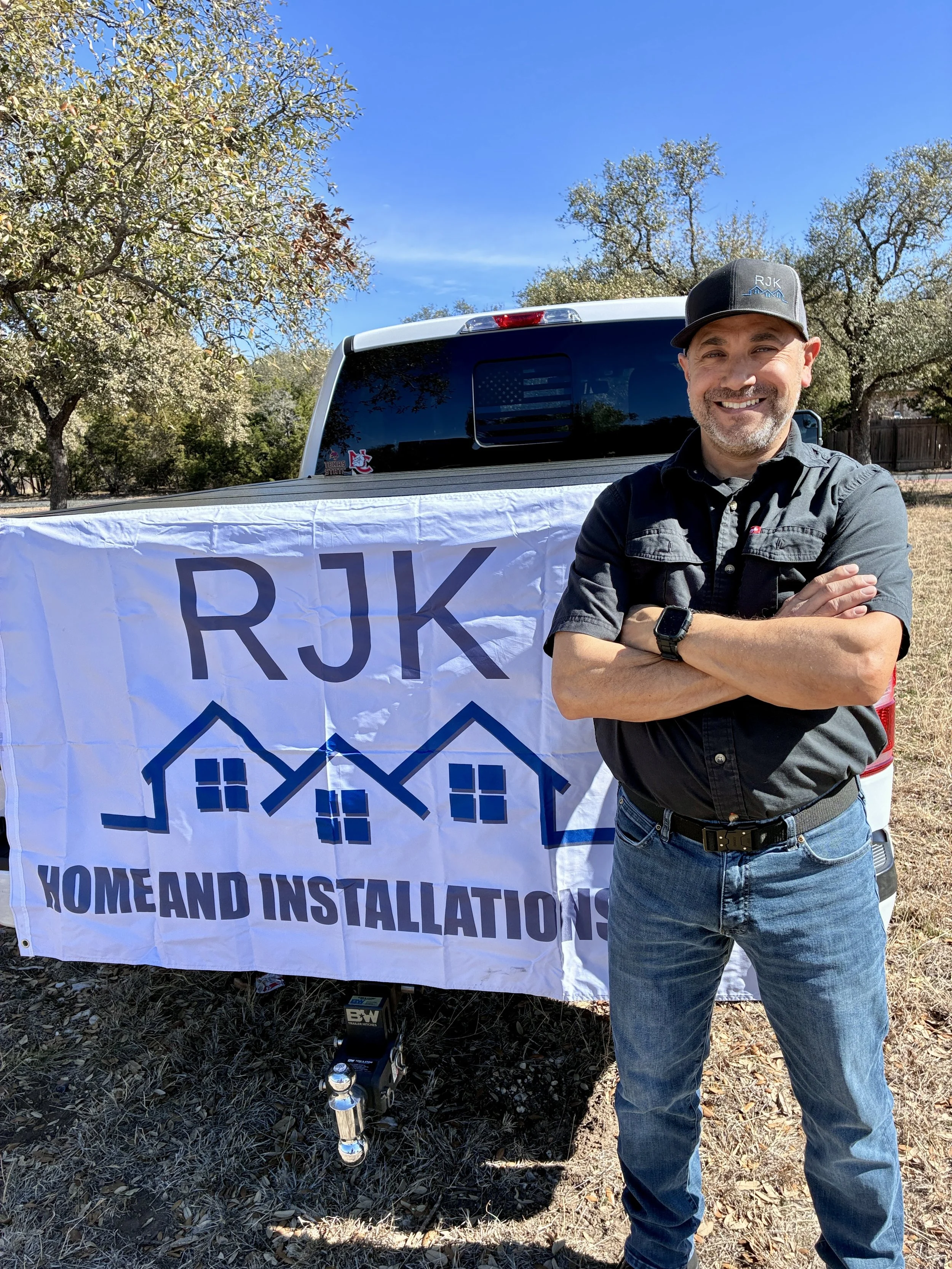 Man standing outdoors smiling, next to a large banner that reads 'RJK HOME AND INSTALLATION,' with a pickup truck in the background and trees under a blue sky.