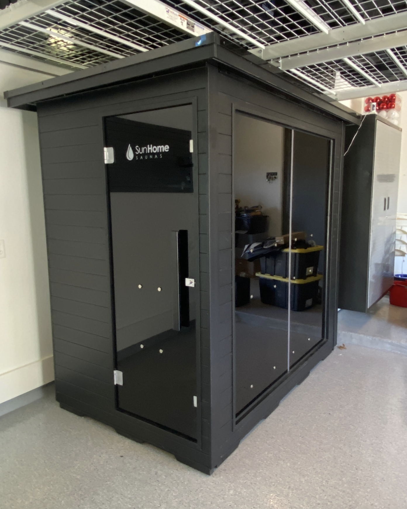 A black sauna unit with a door labeled "SunHome Saunas" is situated indoors on a gray carpeted floor, with ceiling grid and some storage boxes visible inside the sauna.