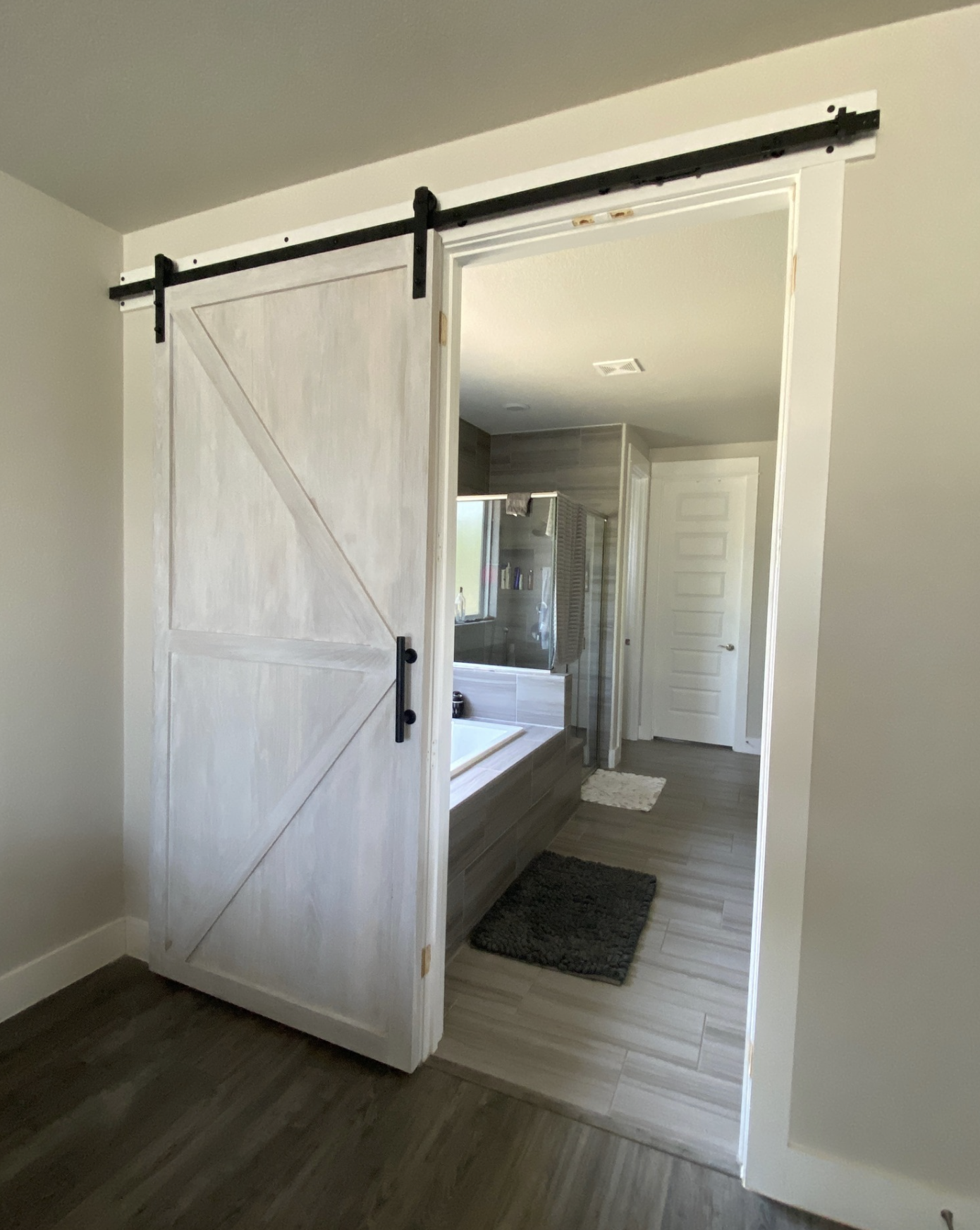 Bathroom with sliding barn door, bathtub, and shower with glass door, gray walls, and wooden flooring.