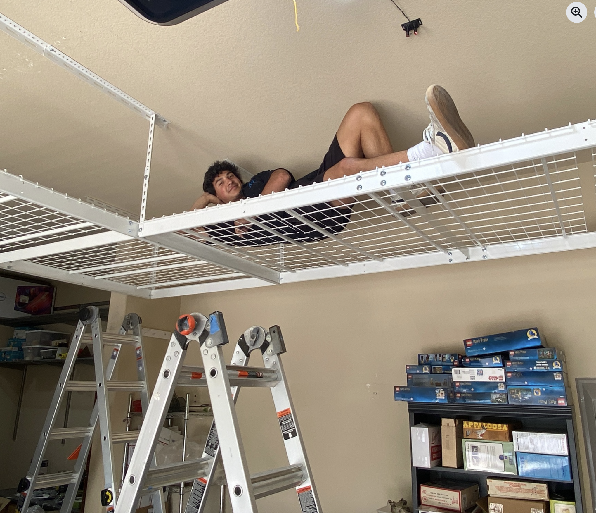 Person lying on a metal wire shelf in a hardware or toy store, with a ladder in the foreground and shelves of toys and boxes in the background.