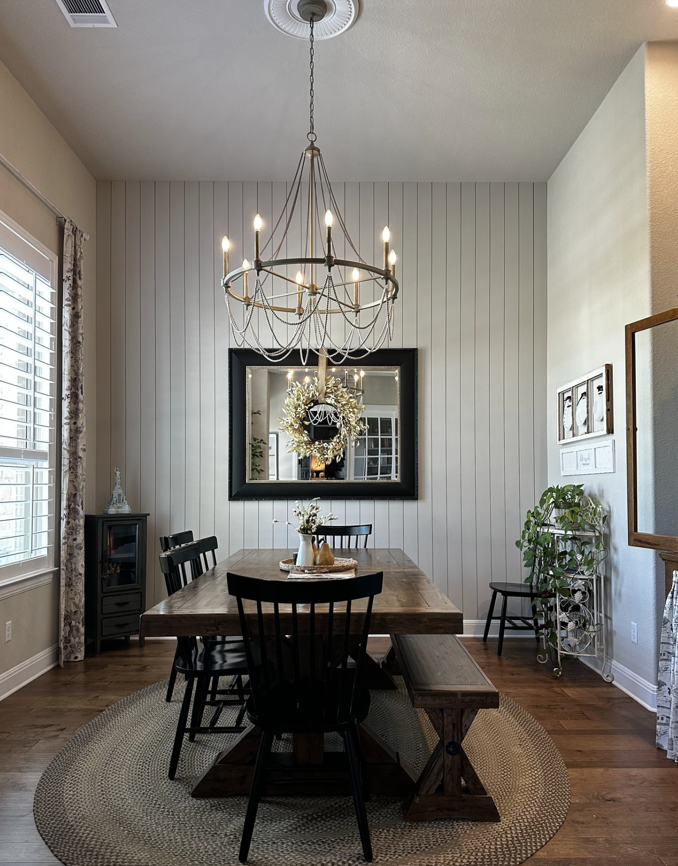 A dining room with a wooden table, black chairs, and a chandelier with candle-shaped bulbs hanging above. A large mirror with a wreath and a vase with flowers sit on the table. There is a window with curtains and a small cabinet with decorative items. The room has wood flooring and a round woven rug under the table.