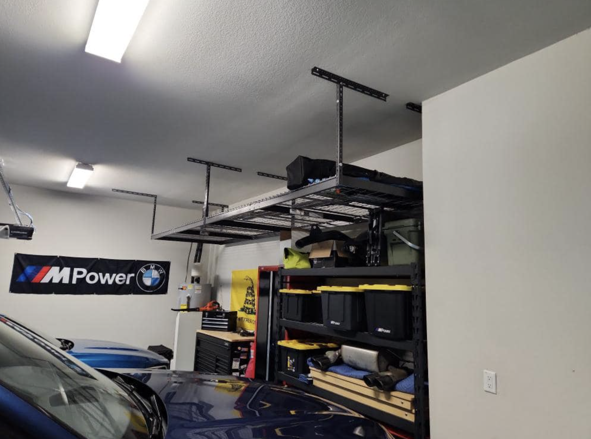 Image of a garage with a BMW M Power banner on the wall, shelves with tools and equipment, a partially visible white car in the foreground, and a ceiling-mounted rack.