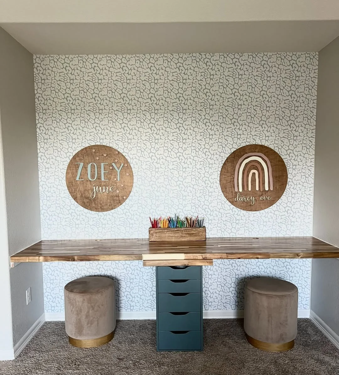 A home office space with a long wooden desk, two round beige stools, and wall decor featuring two round wooden plaques. The left plaque says 'ZOEY june' with small stars, and the right plaque has a rainbow image with the name 'darcy eve.' There is a box of colored markers on the desk.