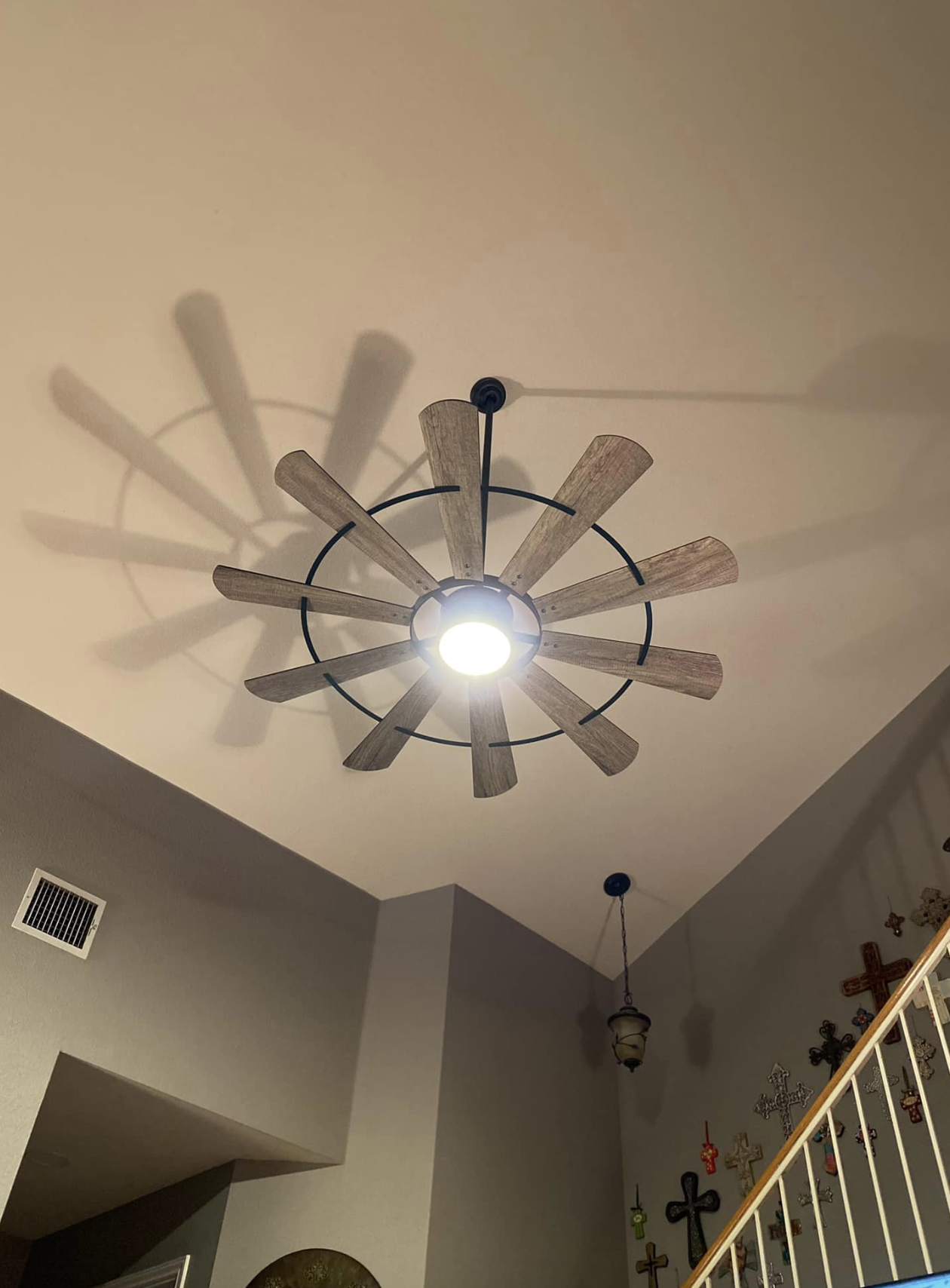 Ceiling fan with wooden blades and central light in a living room with hanging lamp, cross wall decorations, and beige walls.