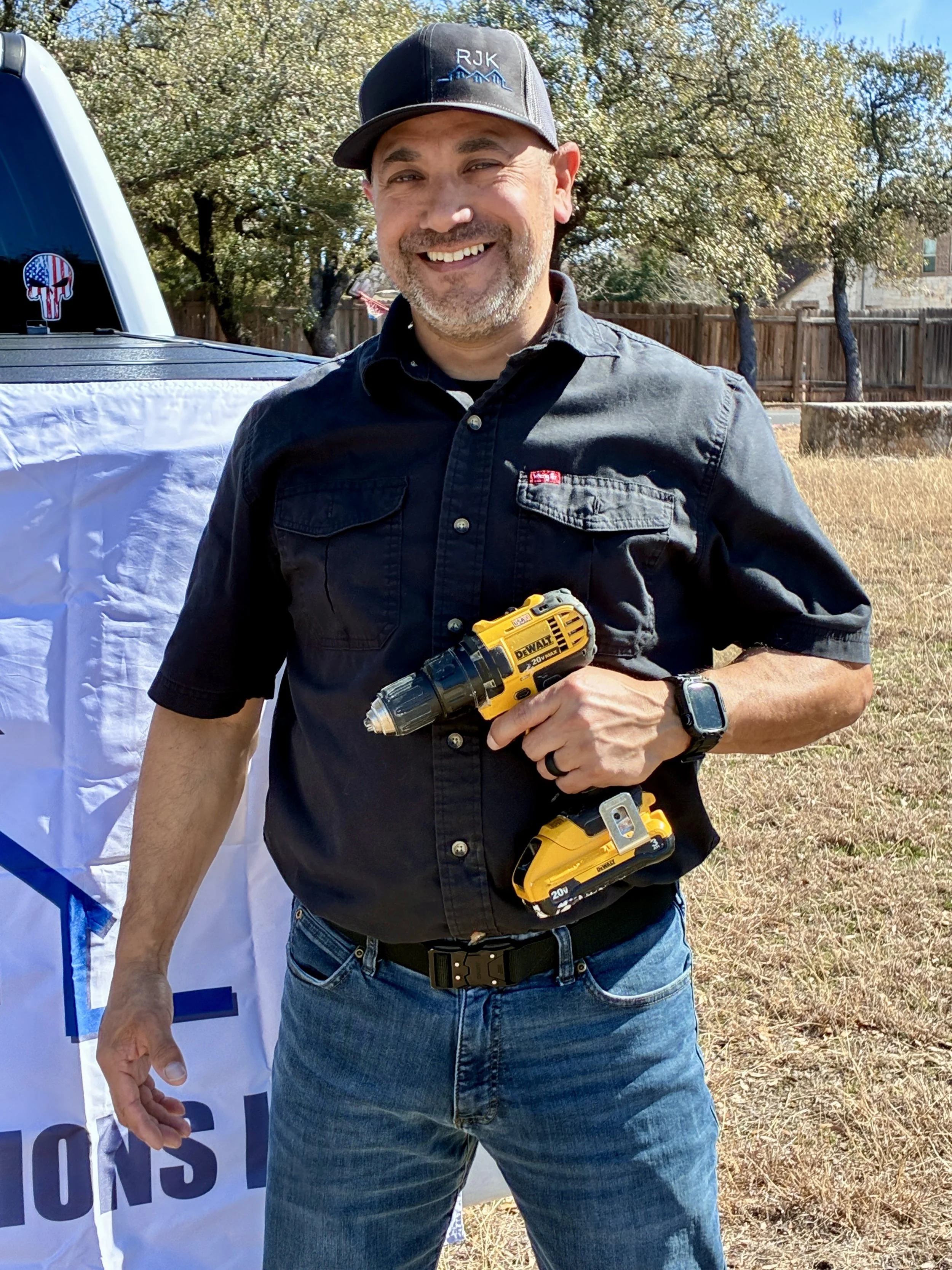 Smiling man in a black shirt and jeans holding a yellow power drill outdoors with trees and a wooden fence in the background.