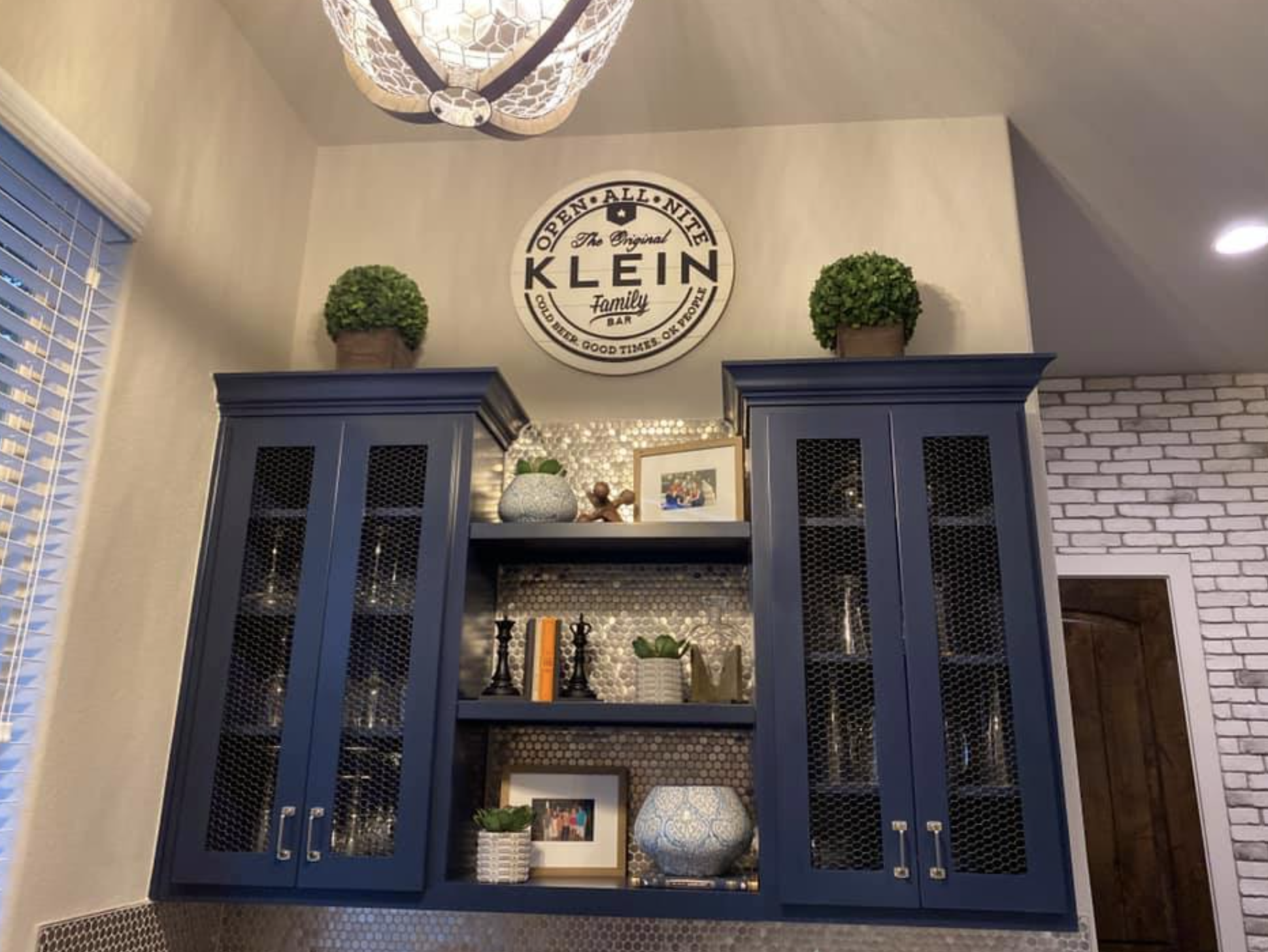 A dark blue wooden china cabinet with glass panel doors, decorated with plants and framed photos on its shelves. Above the cabinet, a round sign reads 'Klein' with additional text, and two potted plants are positioned on top. To the left, there is a window with white blinds, and to the right, part of a brick wall with a doorway.