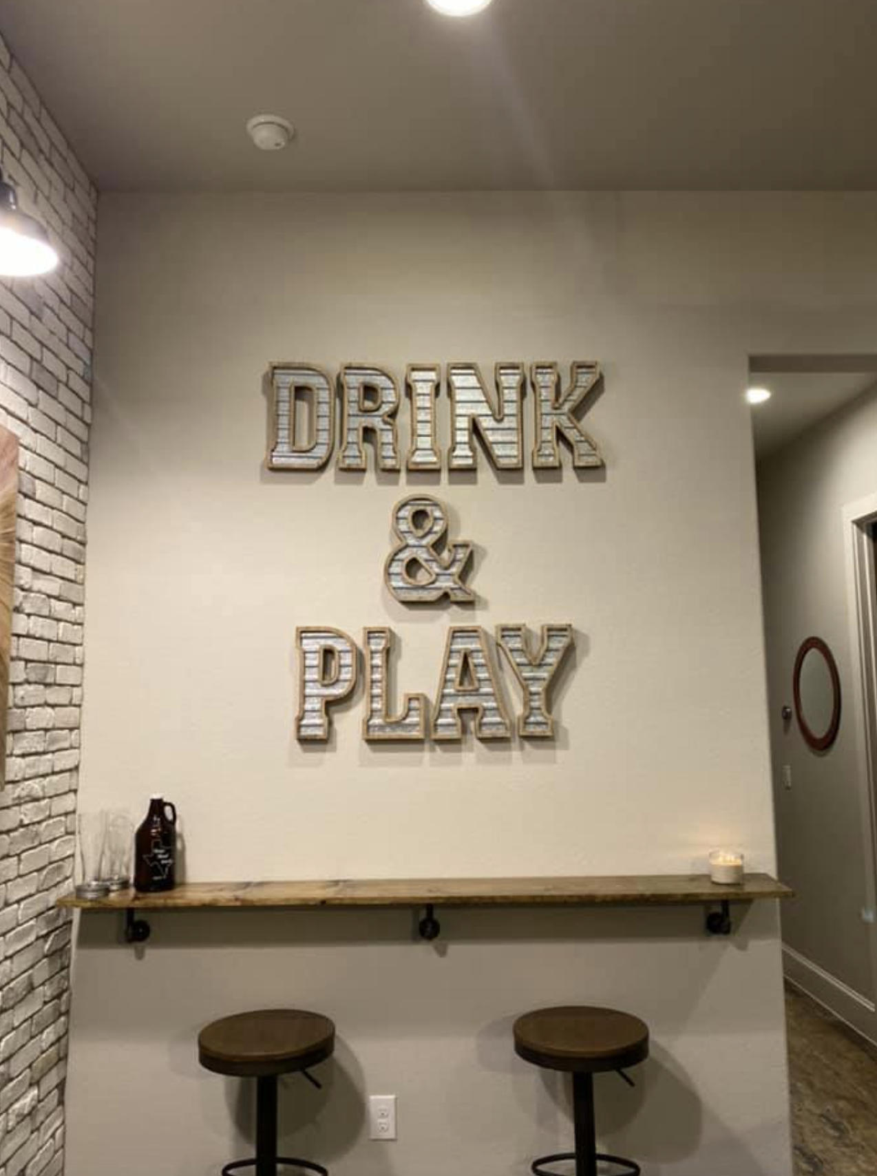 Decorative wall sign that reads 'Drink & Play' in metallic letters, situated above a small bar table with two stools, in a cozy interior space.