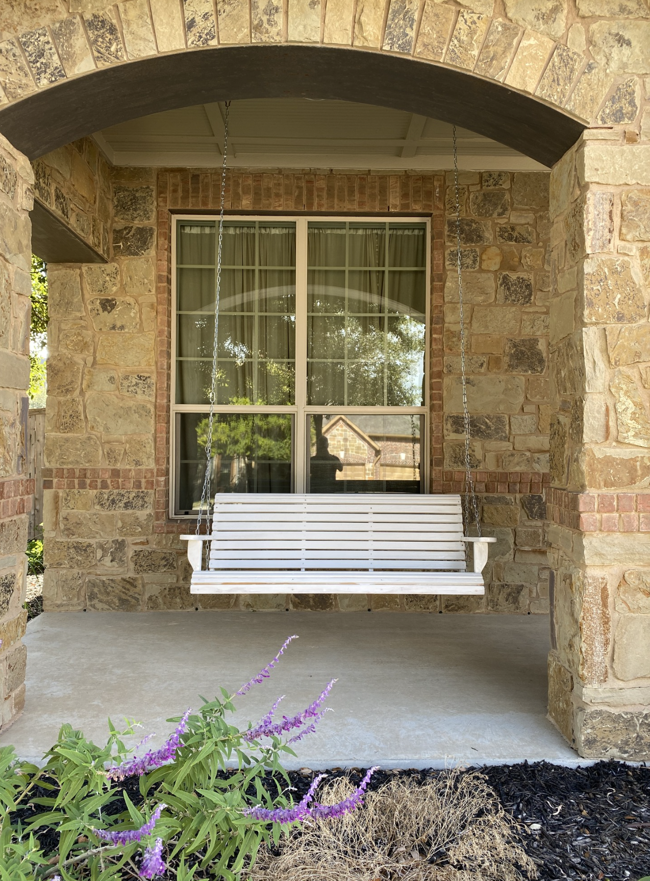 A white porch swing hanging from chains in front of a large window with a brick and stone house exterior. Purple flowers and some dried plants are in the foreground.