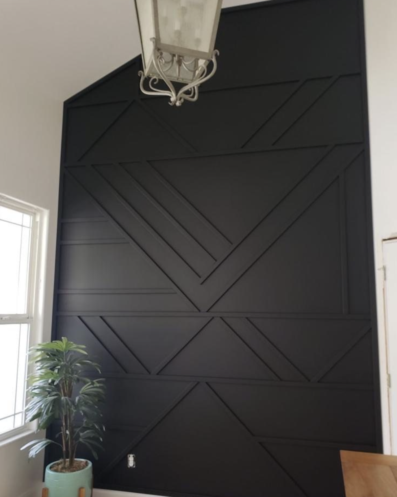 Interior of a room with a black geometric accent wall, a window with white trim on the left, a potted plant on the left, a wooden table at the bottom right, a white metal chandelier hanging from the ceiling, and a partially visible white door on the right.